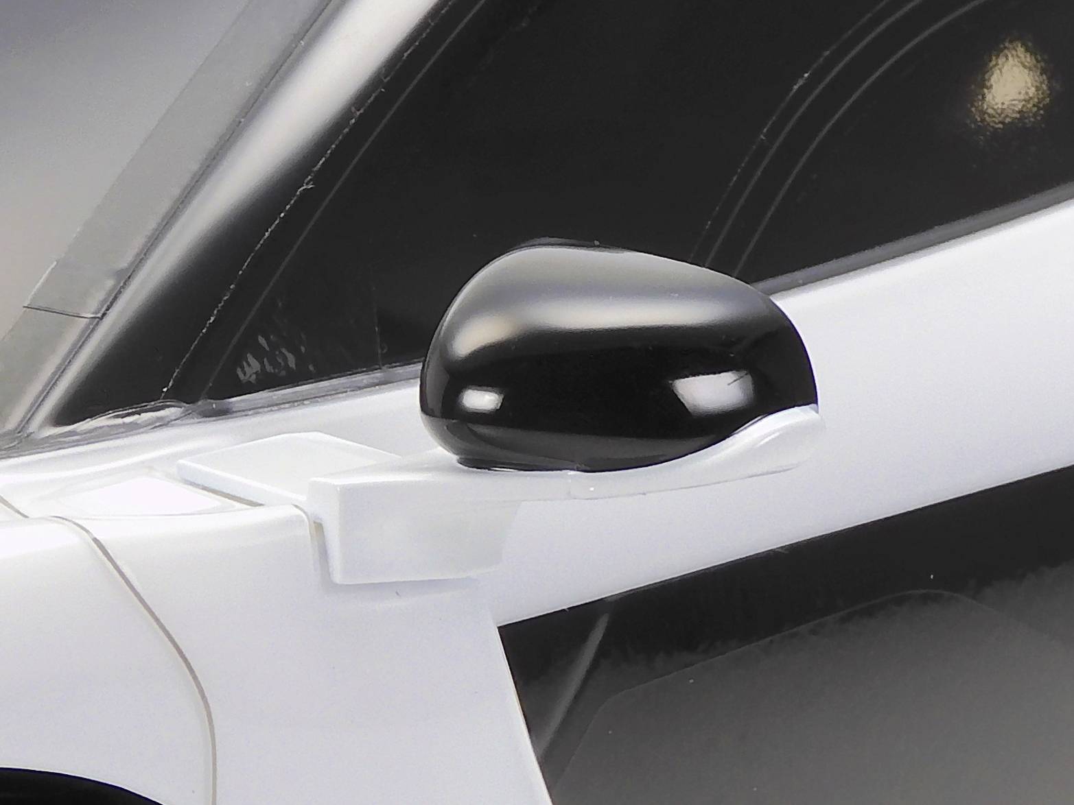Bliski kadra crnog retrovizora na bijelom automobilu, pri čemu su vidljivi detalji karoserije i nosača retrovizora.