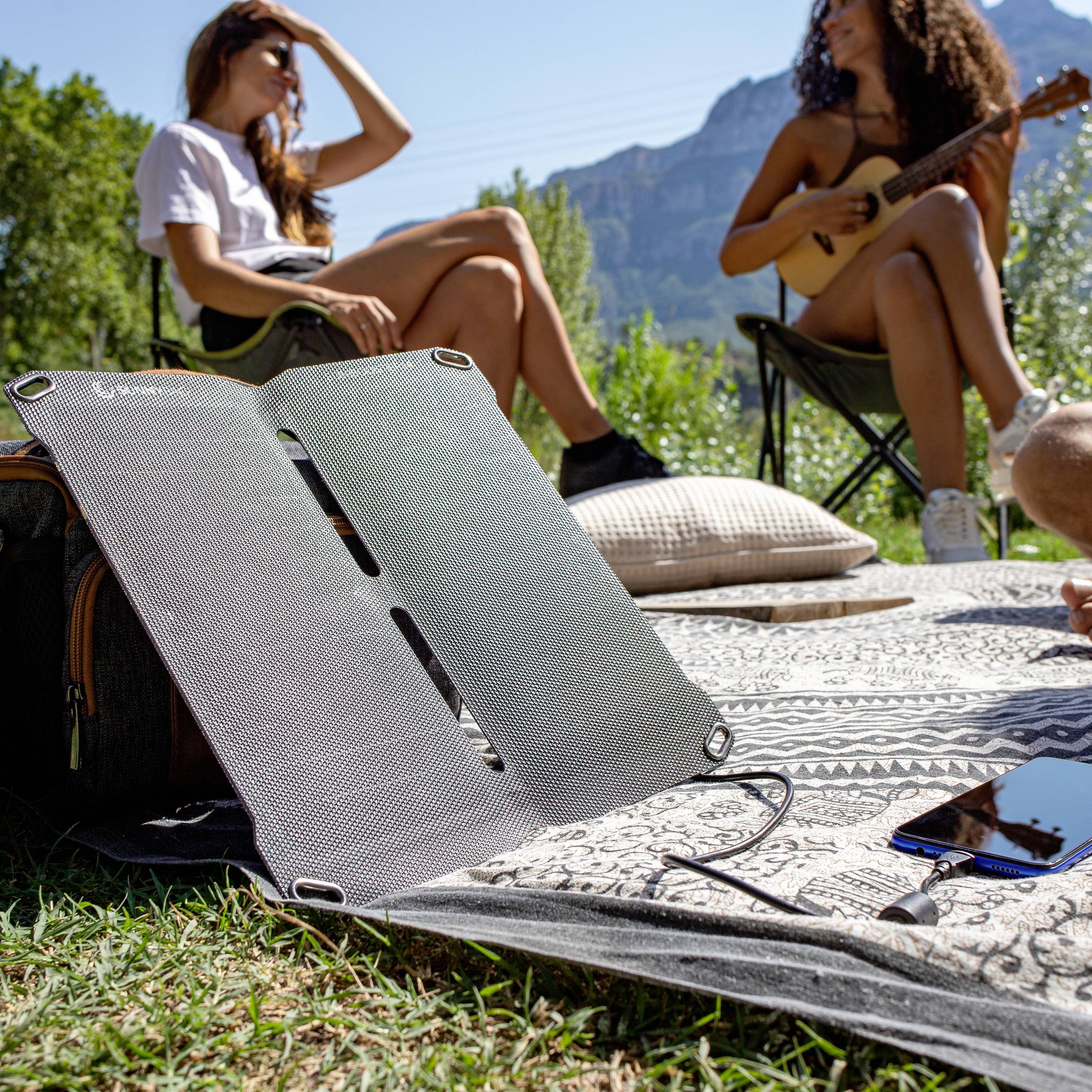 Grupa mladih ljudi opušta se na livadi. Jedna osoba svira gitaru, dok drugi udobno sjede, a solarni panel punjač punи mobitel.