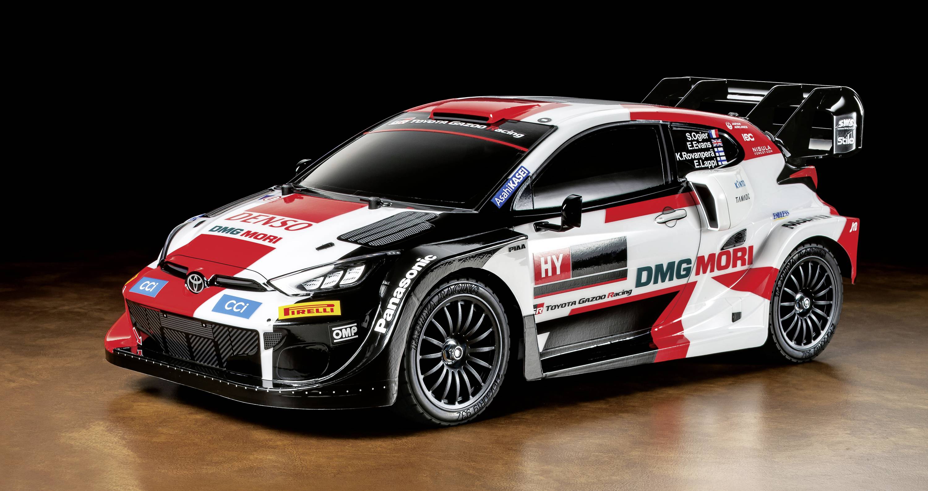 Bijelo-crveni rally automobil sa sponzorskim logotipovima i uočljivim aerodinamičkim elementima stoji s bočne strane na smeđoj podlozi pred crnom pozadinom.