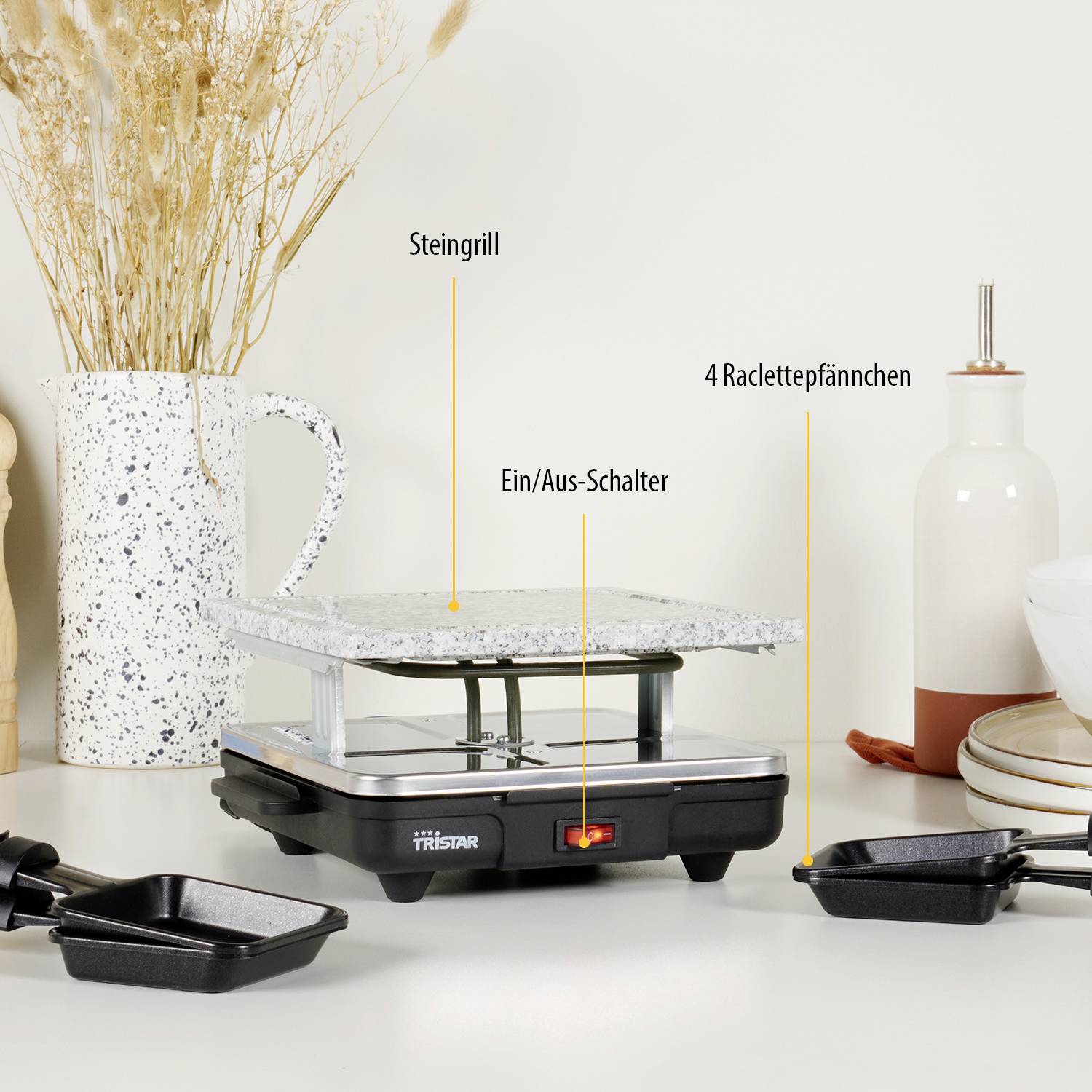 Kameni raclette roštilj na bijelom stolu, označen natpisima 'Steingrill', 'Uključivač/isključivač' i '4 raclette posudice', okružen dekorom.
