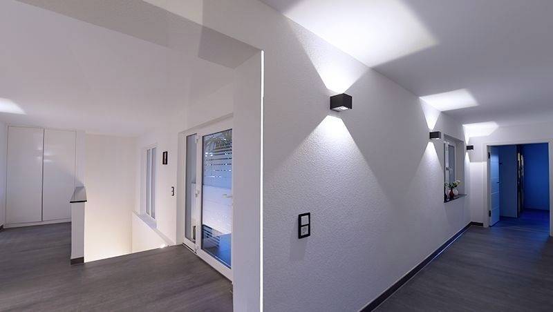 Moderni, minimalistički hodnik s tamnim podom i bijelim zidovima, osvjetljen zidnim lampama; lijevo velik ogledalo.