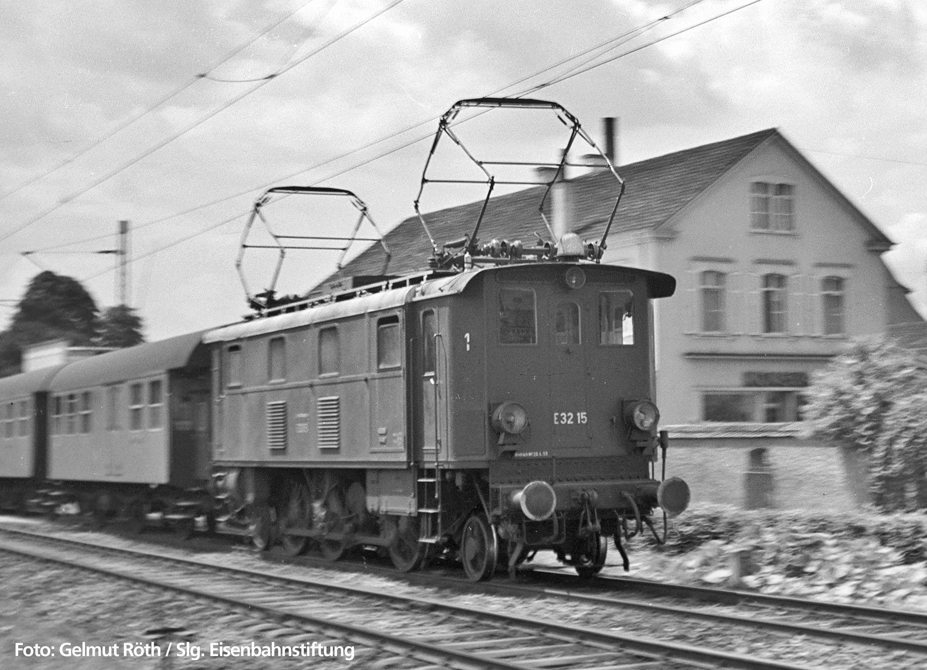 Crno-bijela fotografija povijesne električne lokomotive E 32 15 koja vozi pokraj putničkog vagona ispred zgrade.