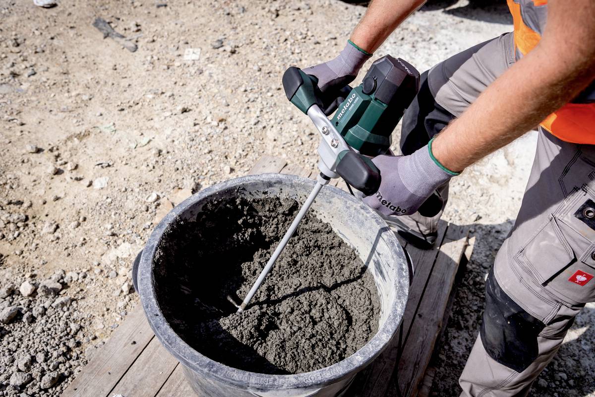 Jedna osoba miješa cement u kanti električnom mješalicom. Nosi radne rukavice i narančasti zaštitni kombinezon.