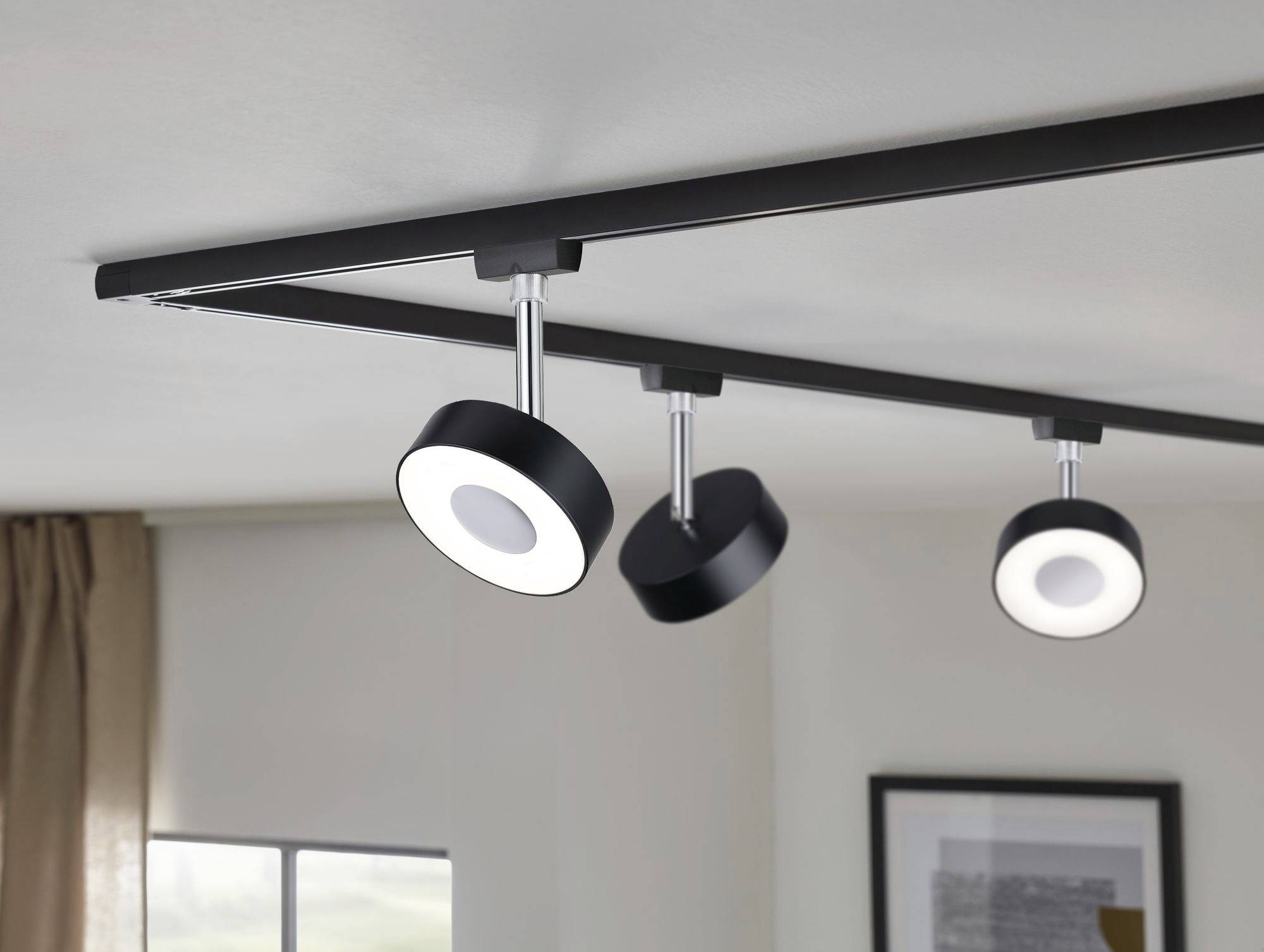 Crni sustav šina s tri okrugle, podesive stropne lampe u modernom prostoru, djelomično vidljivi prozor i slika.