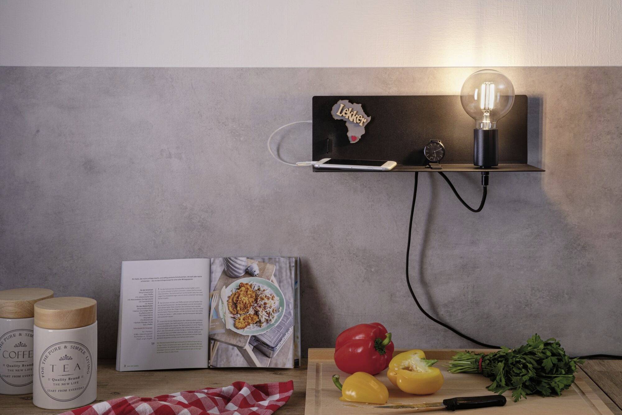 Otvorena kuharska knjiga prikazuje jelo od tjestenine pored crvenih i žutih paprika, peršina, lampe i stolne svjetiljke na drvenom stolu.