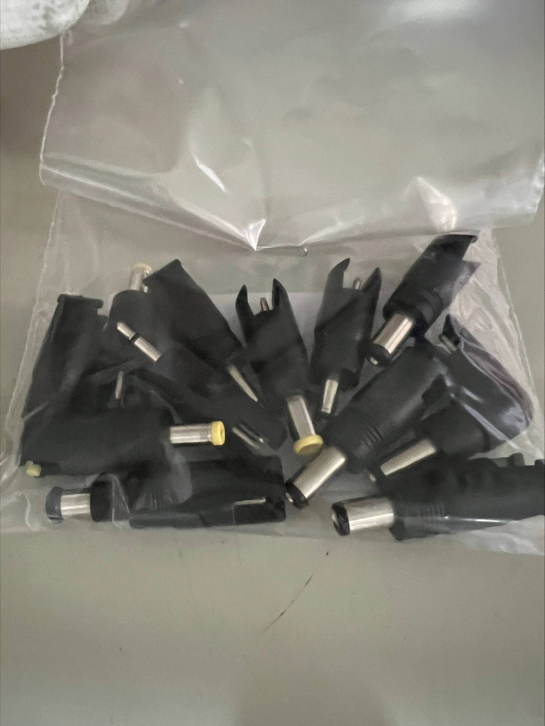 Plastična vrećica sa nekoliko crnih mrežnih adapterskih utikača različitih veličina, nesređeno razasutih po stolu.
