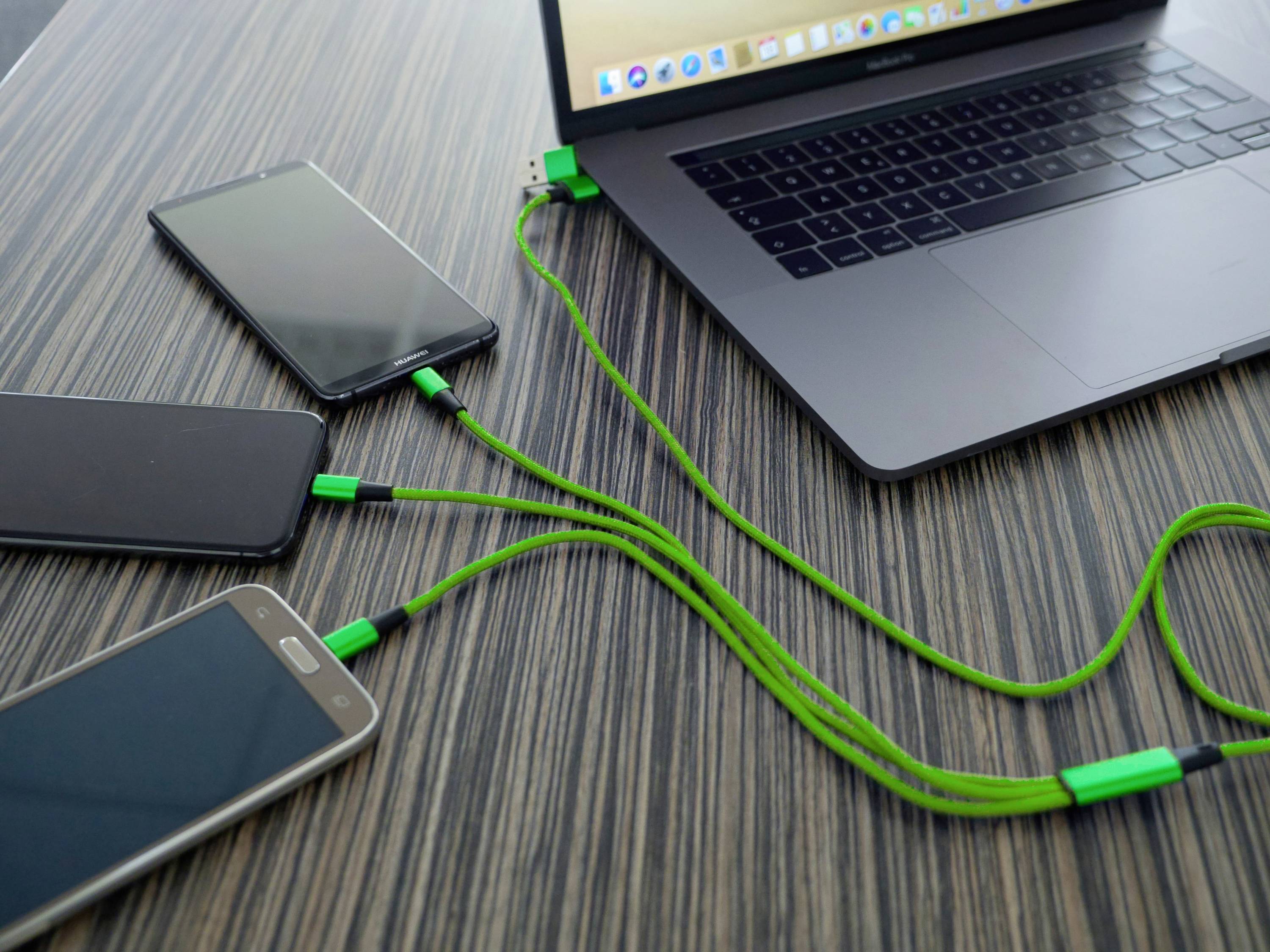 Prijenosno računalo istovremeno punjenje tri pametna telefona putem zelenih USB kabela koji leže na drvenom stolu.