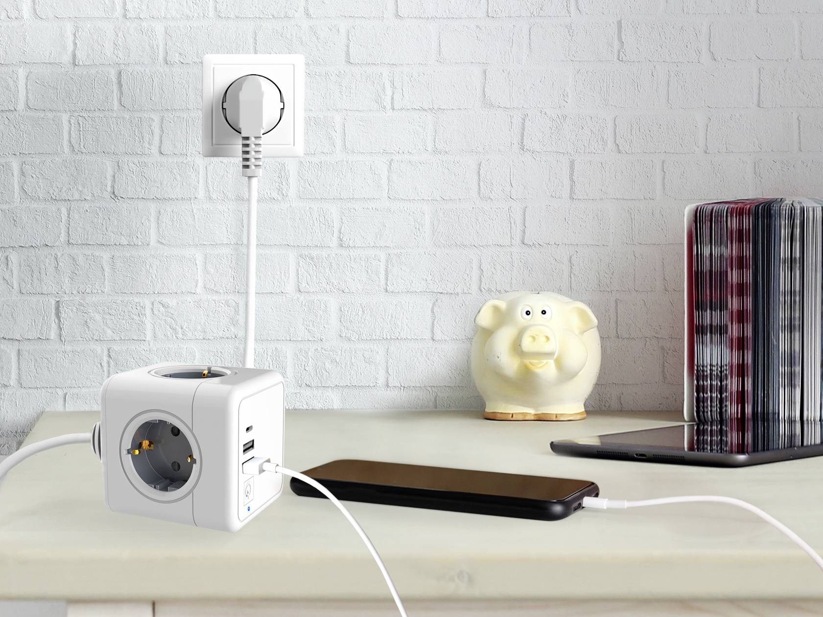 Adapter za utičnice u obliku kocke s dva USB priključka punjač je za pametni telefon. Pokraj njega, na polici, stoji kasica u obliku praseta.