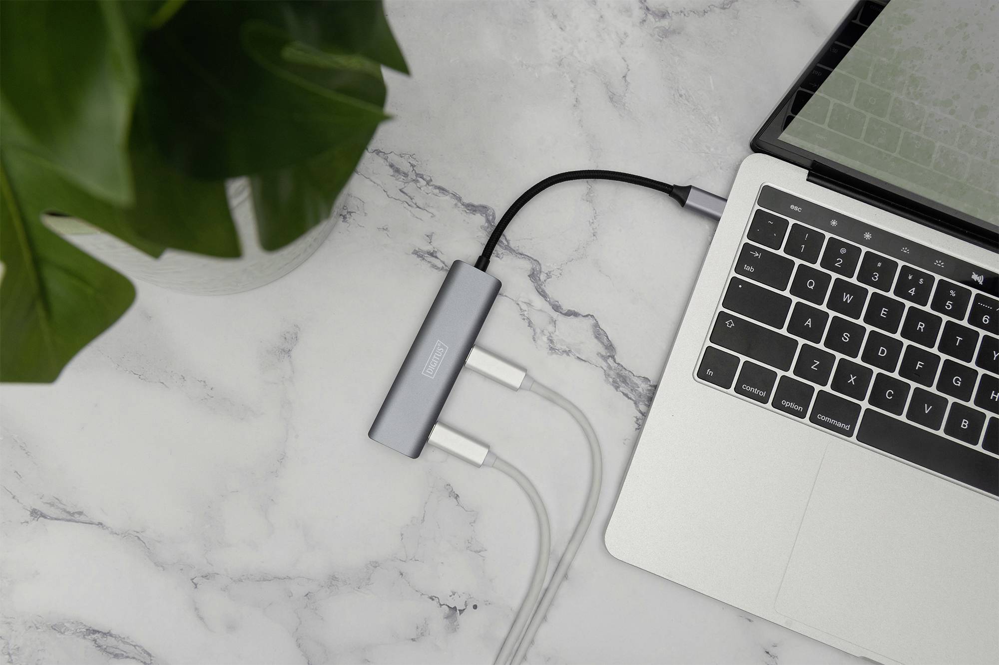 Laptop na mramornoj površini s priključenim USB hubom i kabelima. Lijevo u slici vidljiva je zelena biljka.