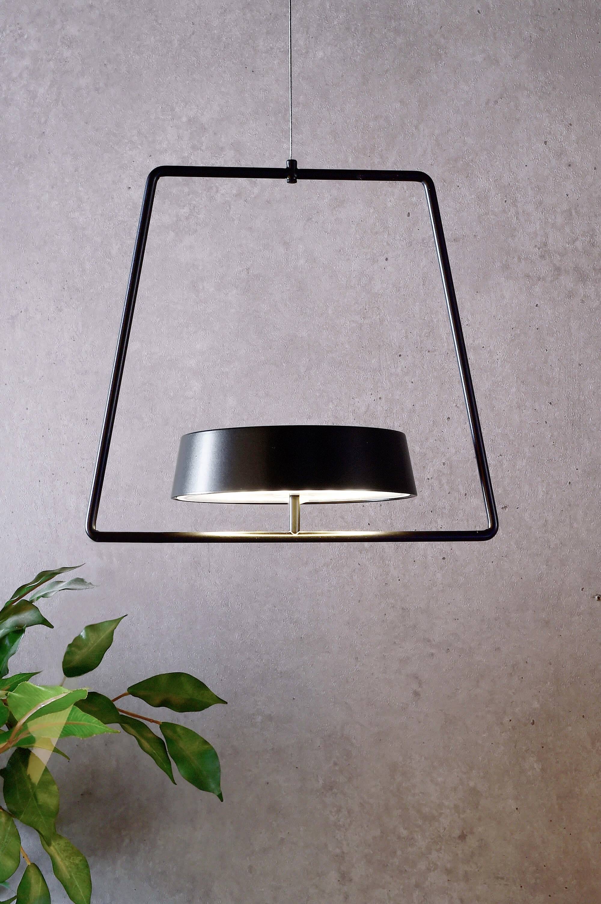Moderna crna viseća lampa u trapezastom okviru ispred sive stijene; lijevo dolje vidljivi zeleni listovi biljke.