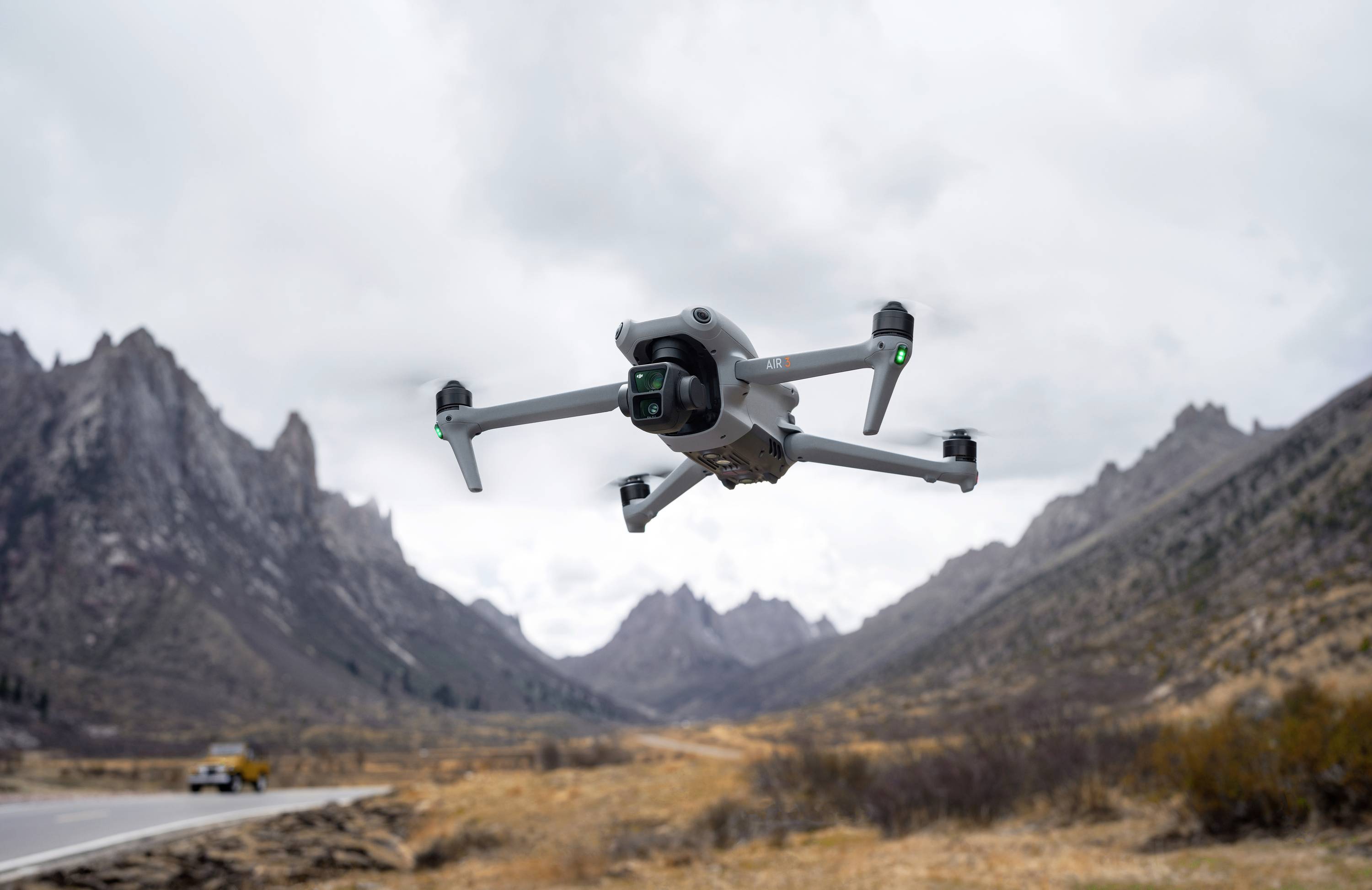 Dron leti iznad planinskog krajolika s oblačnim nebom. U pozadini se naziru strmi vrhovi i automobil na cesti.