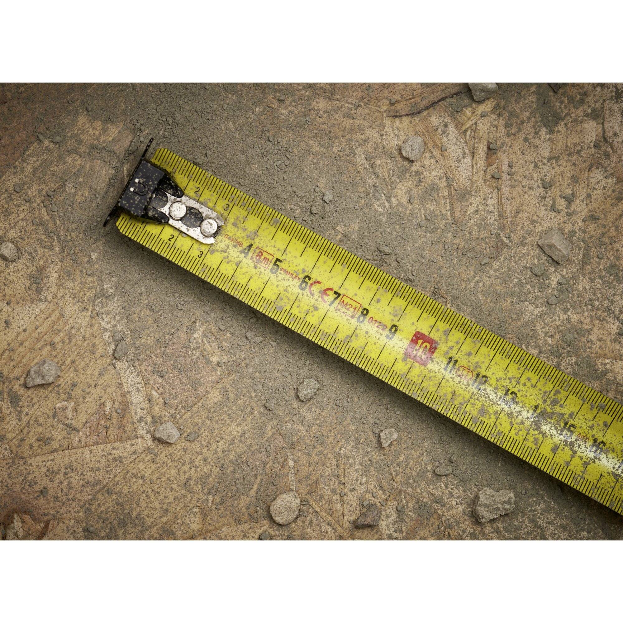 Žuti mjerni trak leži koso na prašnjavom tlu s kamenjem. Prikazuje odsječke u centimetrima i milimetrima.
