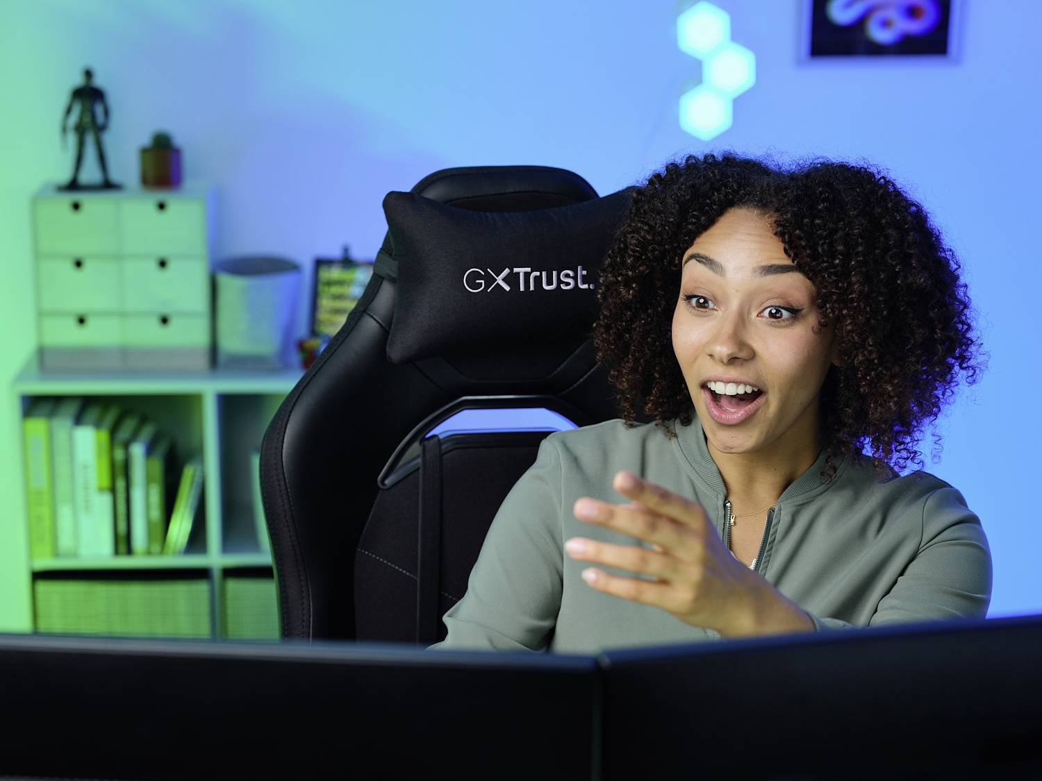 Žena kovrčave kose sjedi u gaming stolici ispred monitora, oduševljenog izraza. Prostorija je osvjetljena plavom rasvjetom.