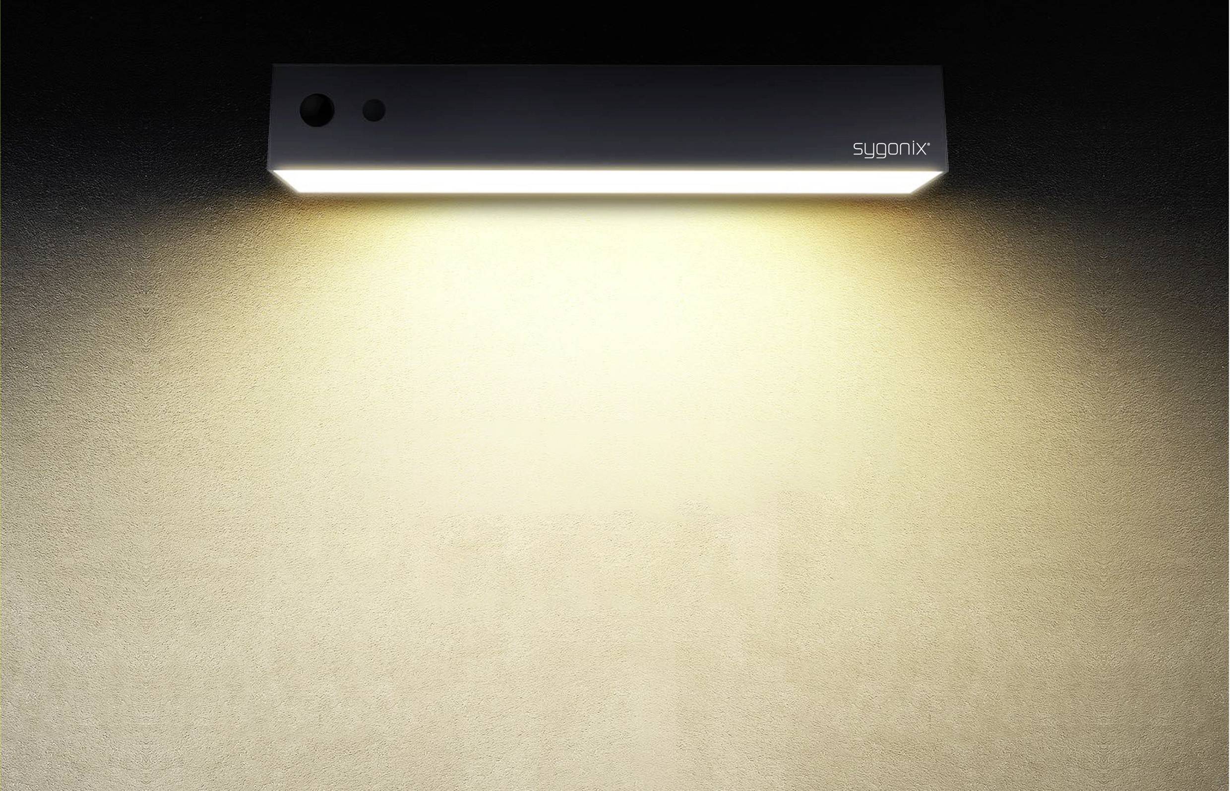 Pravokutna zidna lampa emitira meko, toplo bijelo svjetlo na bež zidu. Naziv marke u gornjem desnom kutu.