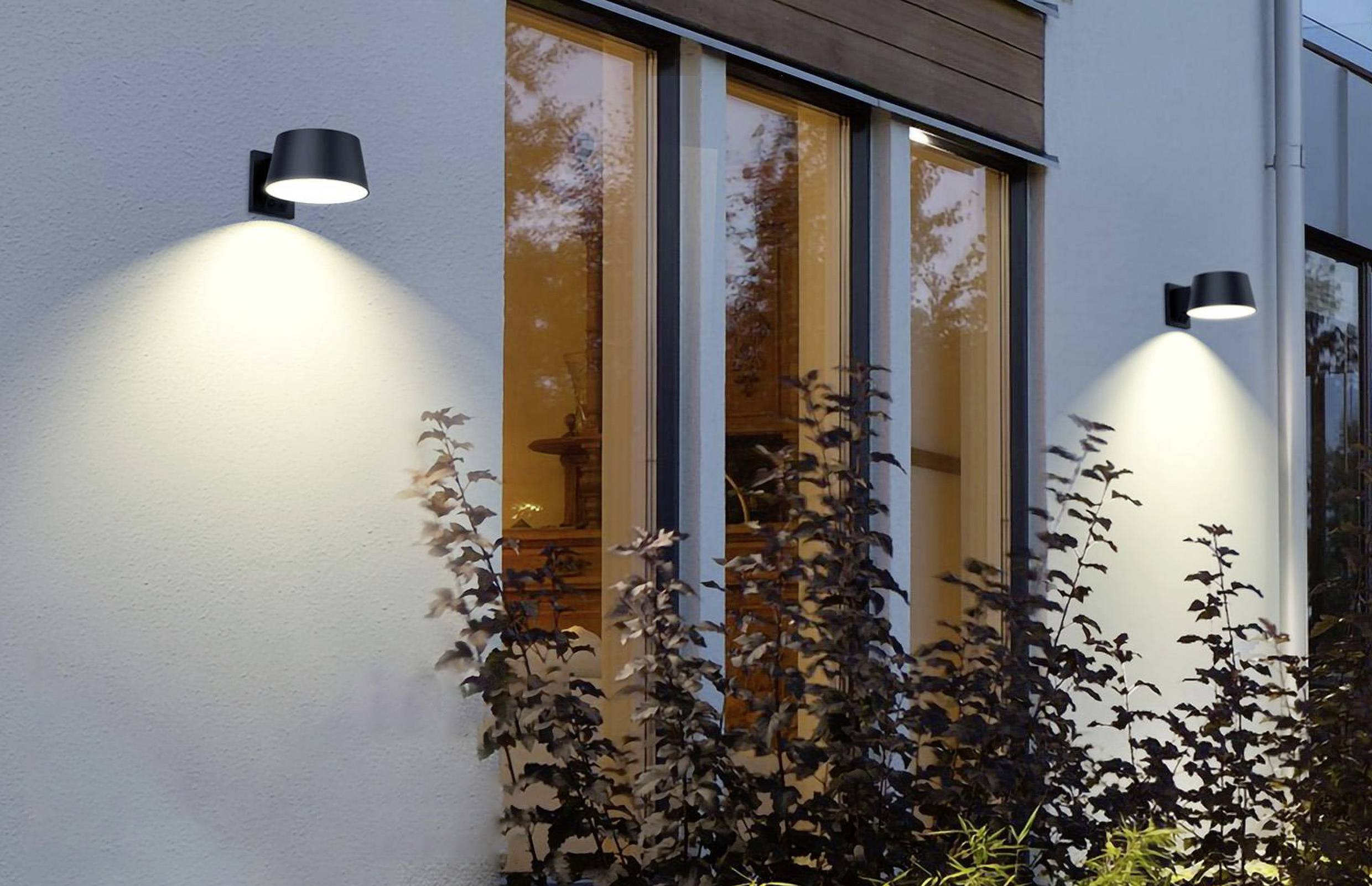 Vanjska rasvjeta na zidu kuće, osvjetljava biljke ispod. Prozori reflektiraju večernje nebo.