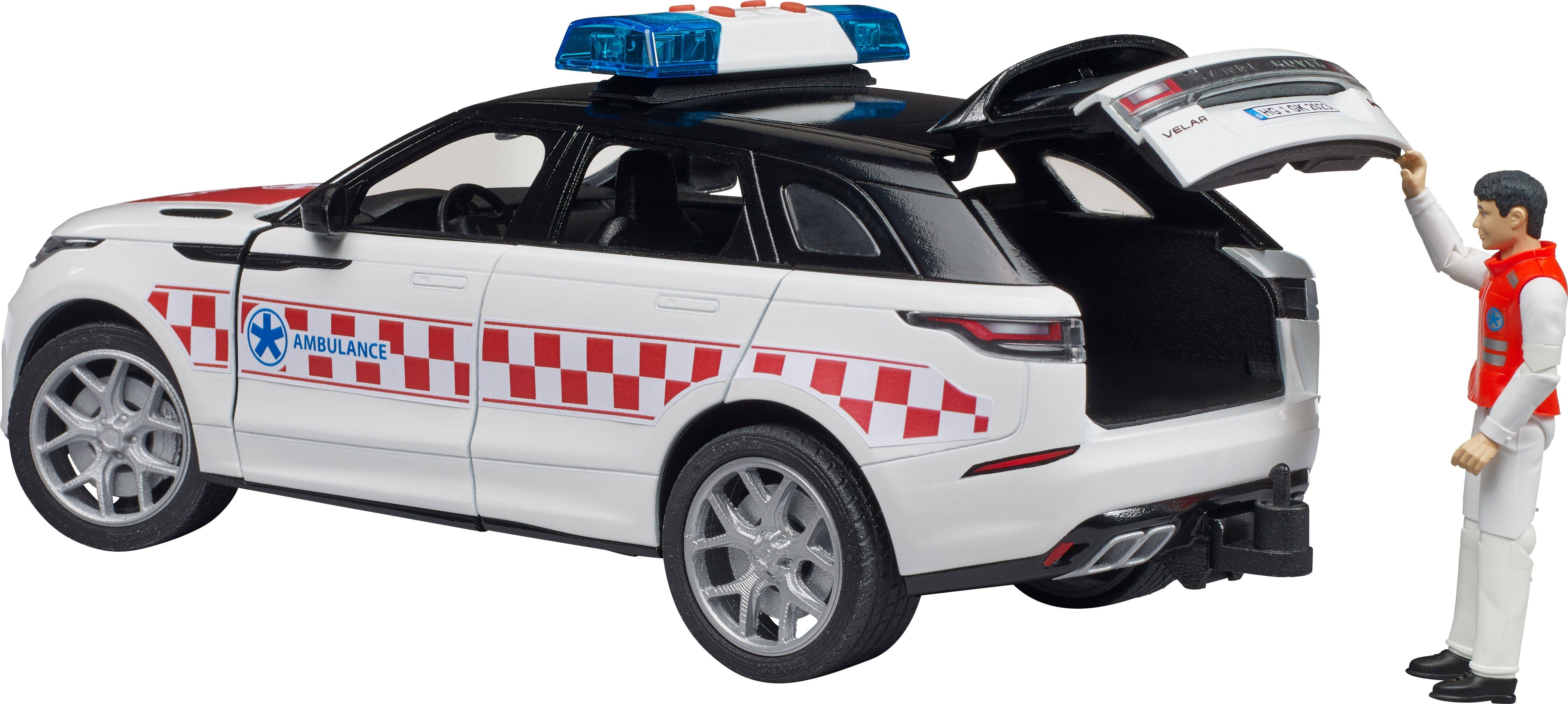 Model igračke hitne pomoći s otvorenim prtljažnikom, pored kojeg stoji figura u crvenoj uniformi koja ga pregledava.