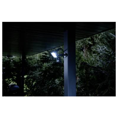 Svijetla verandska lampa montirana ispod tamnog krova pored nosivog stupa, koja osvjetljava šumovit teren noću.