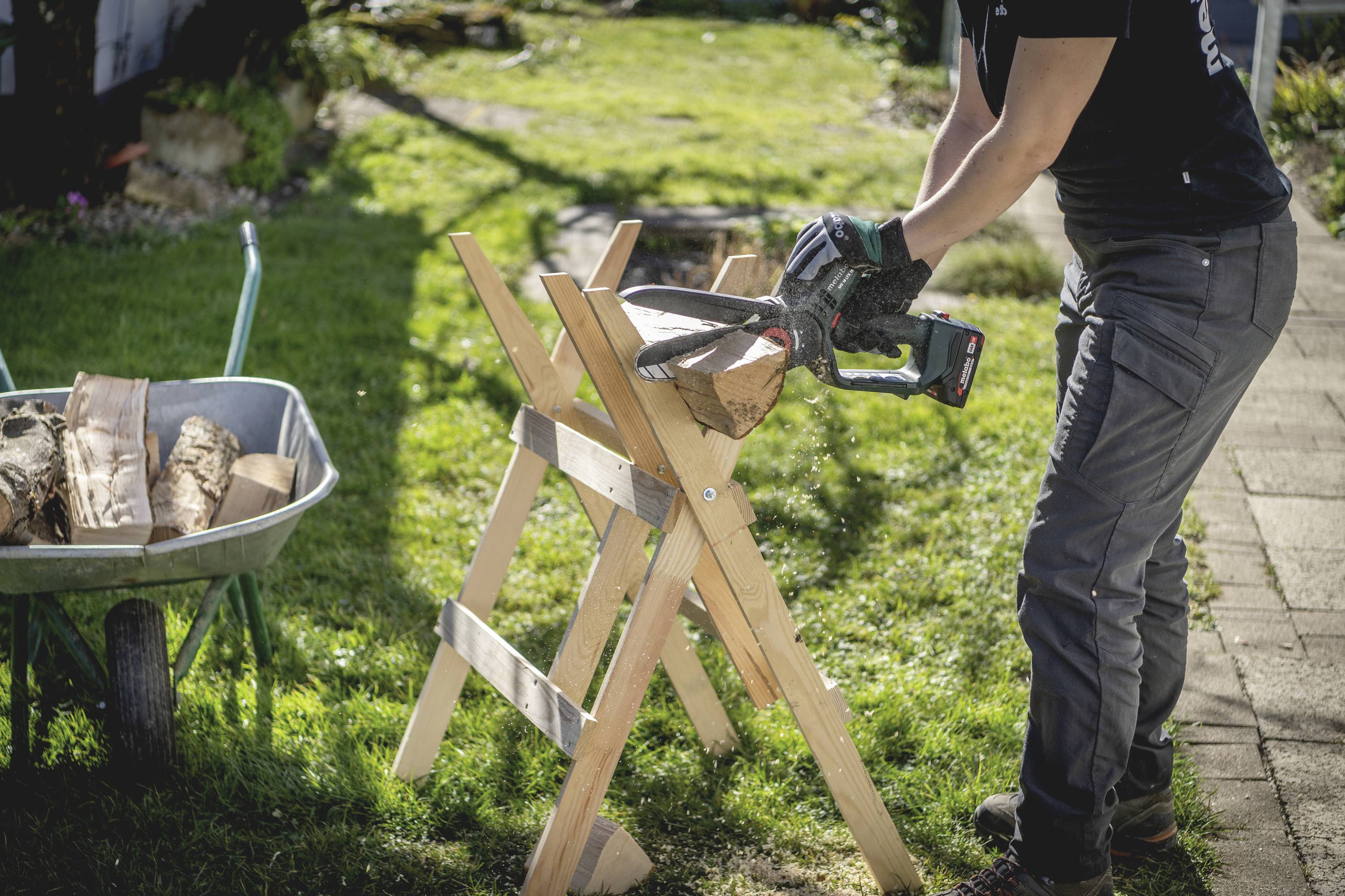 Jedna osoba pile drvo na radnim kobilicama, pored koje se nalazi kolica napunjena drvenim цепаницама.