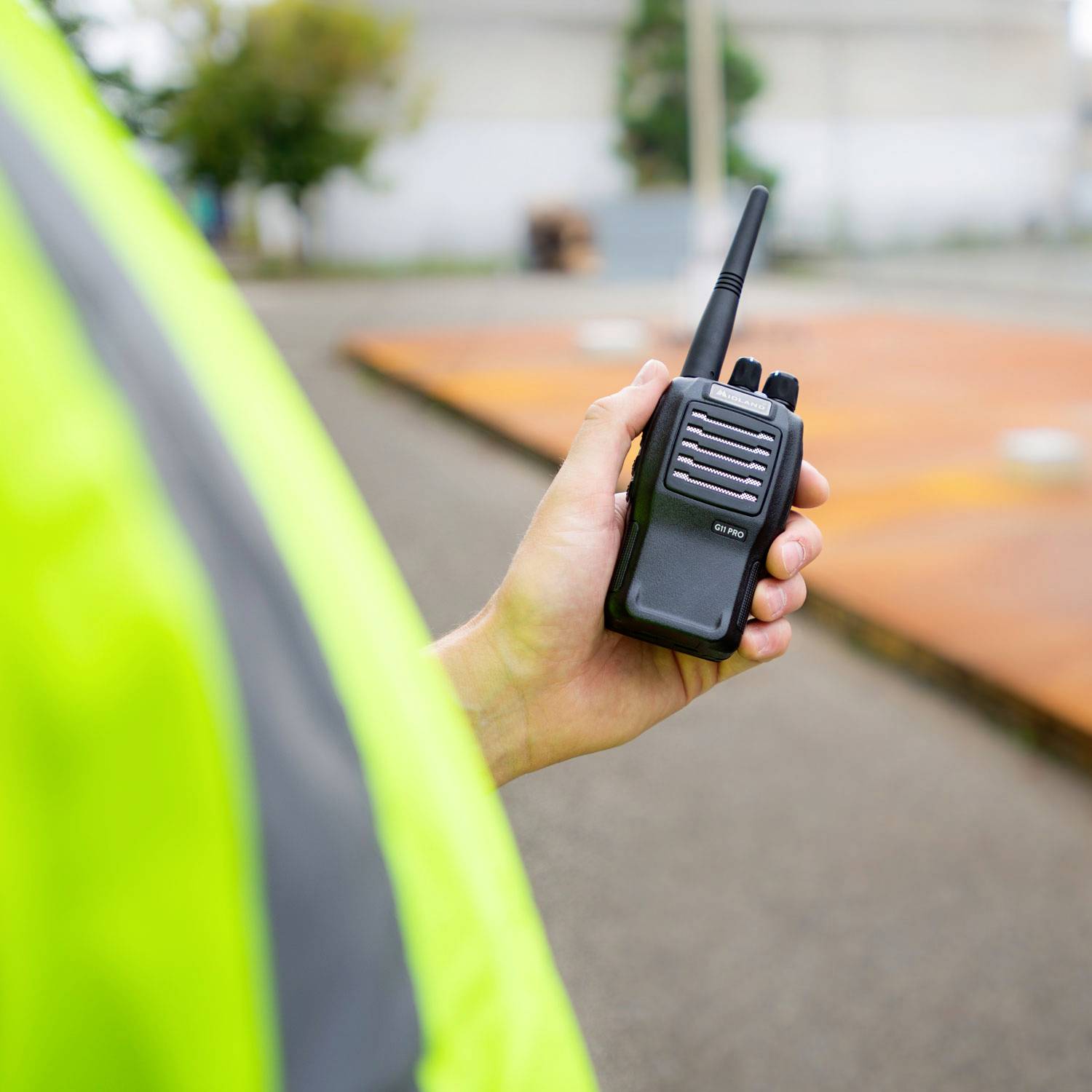 Osoba drži radio u ruci dok nosi neonsko žuti sigurnosni prsluk. Pozadina prikazuje nejasnu cestu.