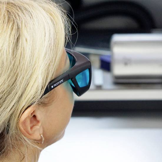Žena nosi naočale za virtualnu stvarnost i promatra nejasnu radnu klupu u pozadini. Njena kosa je plava.
