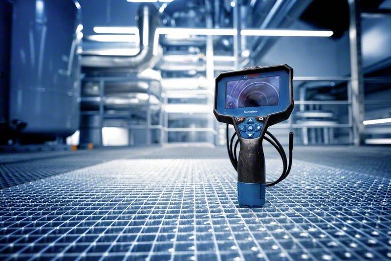 Industrijska okolina s rešetkastim podom, digitalni uređaj za inspekciju postavljen u prednjem planu, metalne cijevi u pozadini.