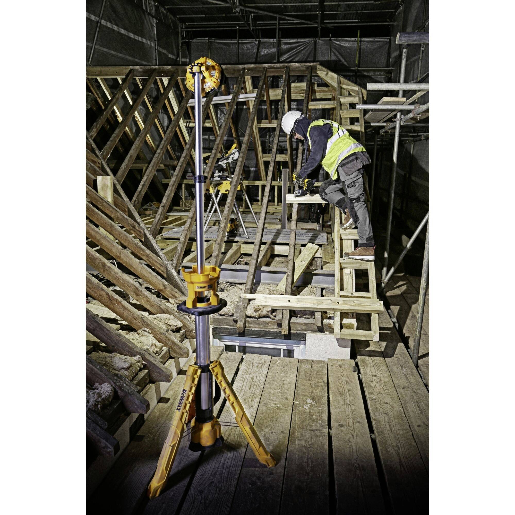 Radnik u zaštitnoj kacigi mjeri drveni okvir na gradilištu, pri čemu se u prednjem planu nalazi stativ s uređajima. Dio gradilišta je prekriven scaffoldingom.