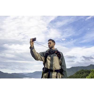 Muškarac u odjeći za vani snima selfie smartfonom ispred pozadine planina i oblačnog neba.