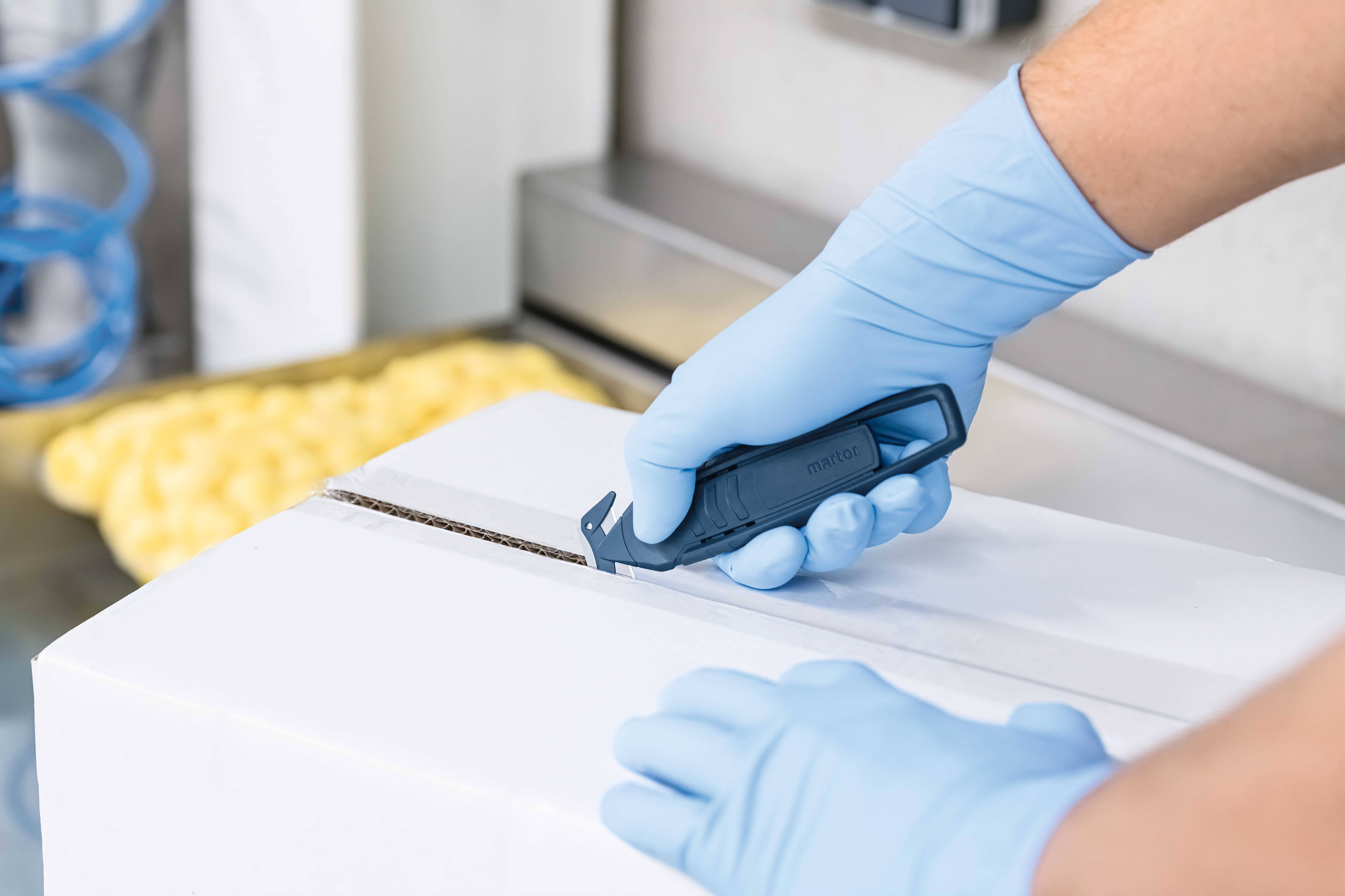 Osoba u plavim rukavicama otvara bijeli karton sigurnosnim nožem u industrijskom okruženju.