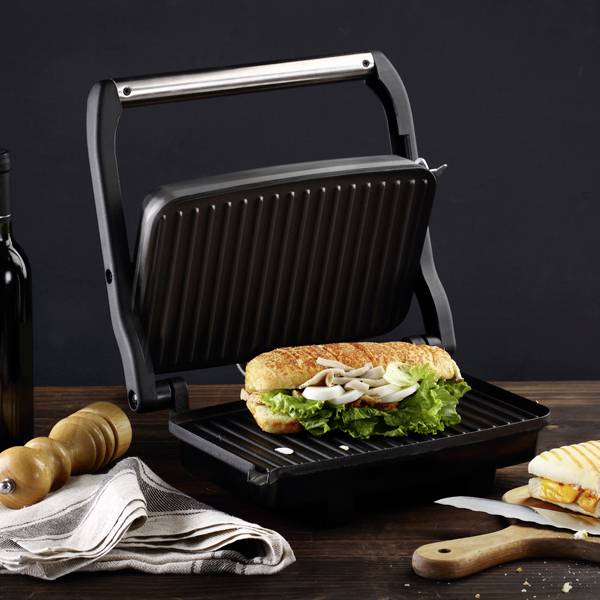 Panini grill pritišće sendvič s salatom i gljivama. Pokraj njega na drvenom stolu leže kuhinjski alati i boca vina.