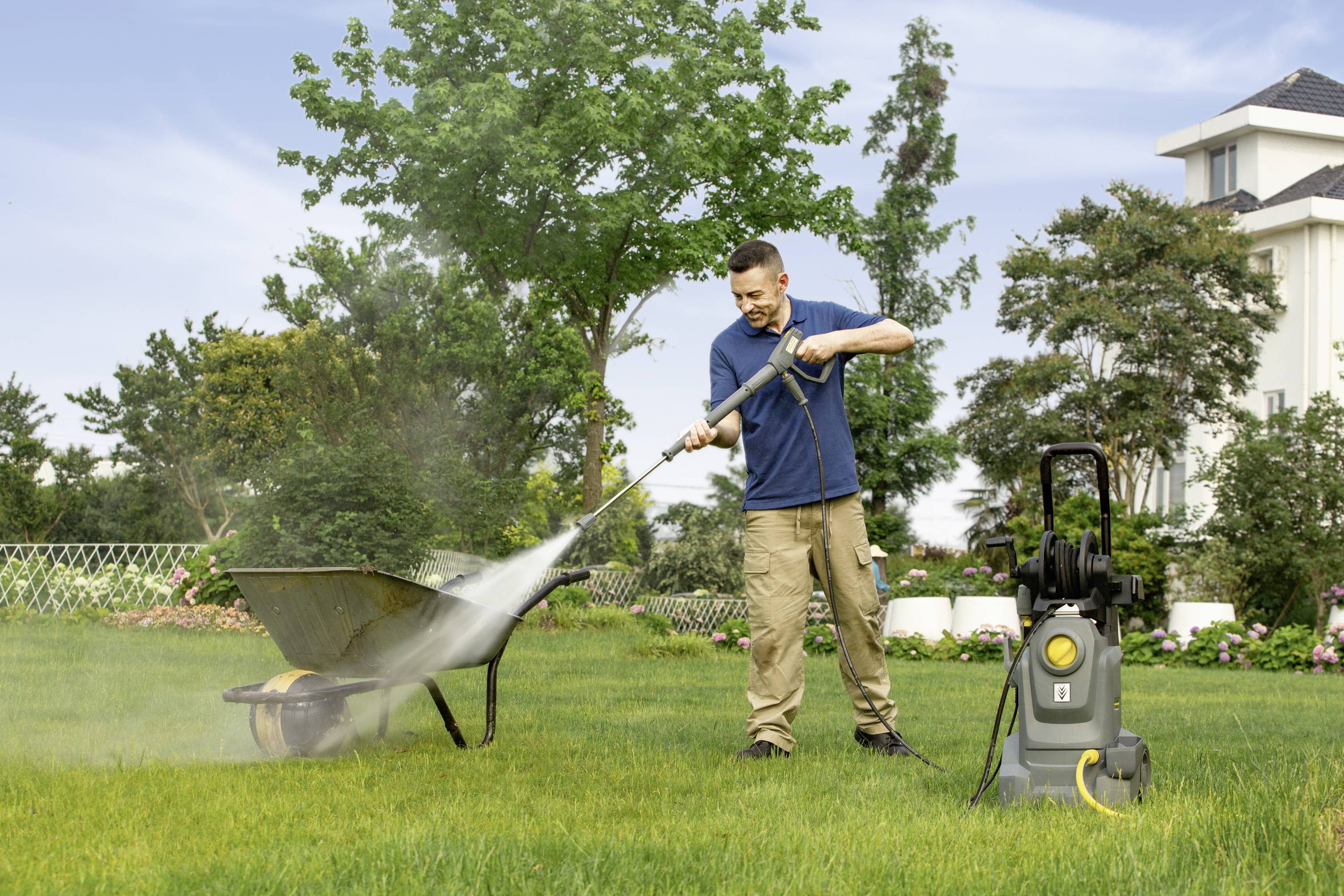 Muškarac čisti tačke visokotlačnim čistačem u vrtu. U pozadini se nalaze stabla i stambena kuća.