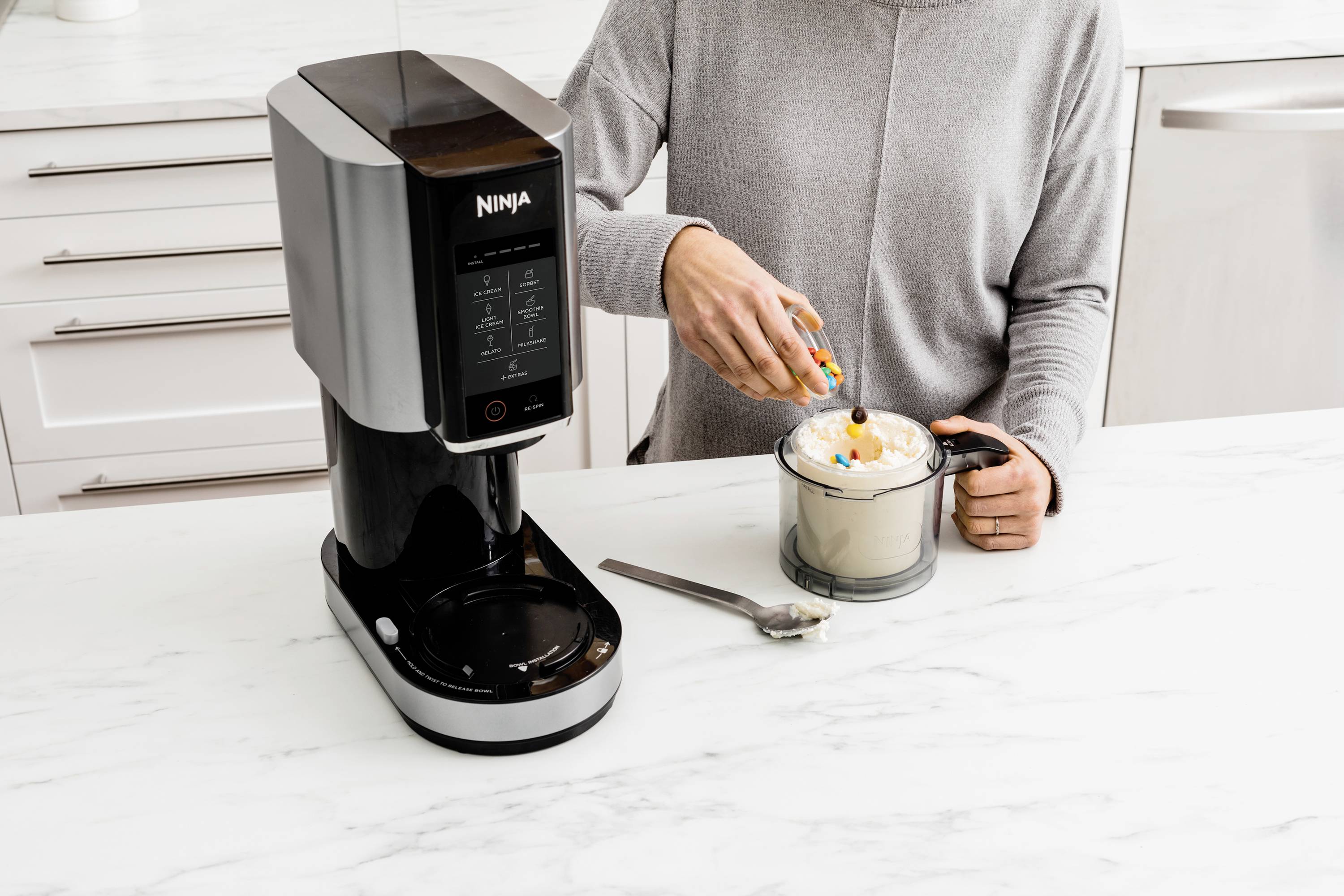 Jedna osoba priprema piće miješalicom za kuhanje, dodajući šlag i šarene posipke na radnoj ploči.