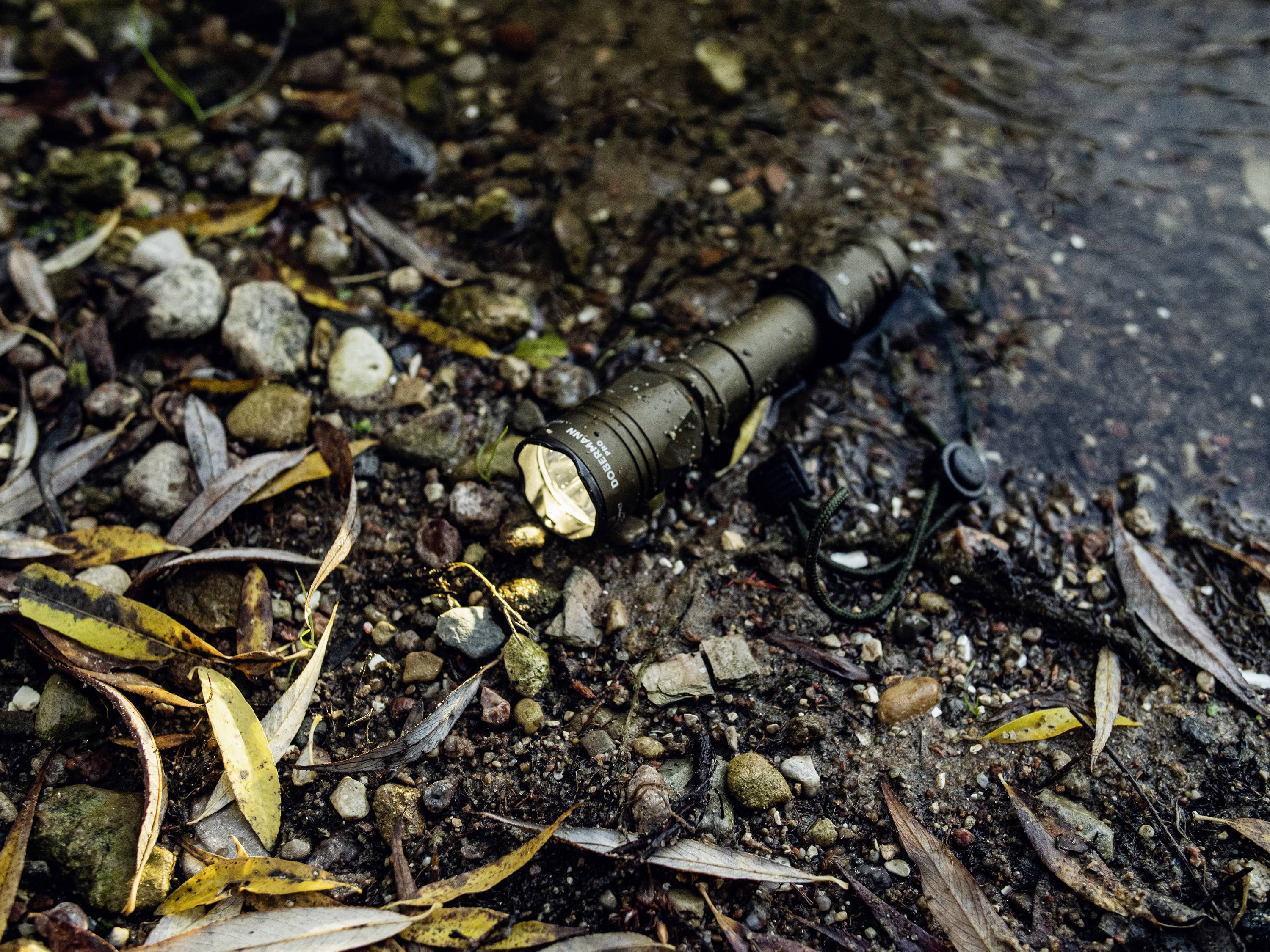 Baterijska svjetiljka leži na obali potoka u šumi, okružena kamenjem i mokrim lišćem. Lampa svijetli unatoč vlazi.