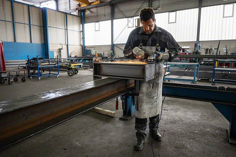 Muškarac radi u radionici, koristi brúsilicu na velikoj metalnoj platformi. Varnice lete okolinom.
