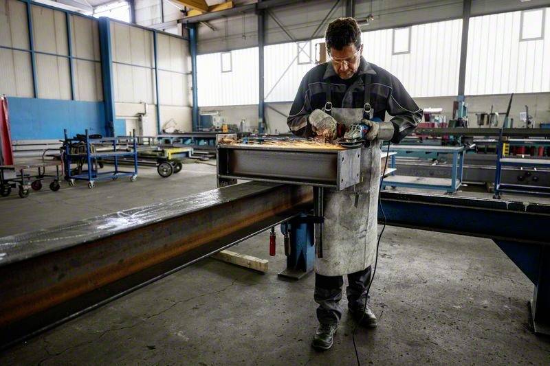 Radnik brusi metal u fabrickoj hali. Nosi zaštitne naočale i pregaču. Iskre lete tijekom brušenja. U pozadini se nalaze strojevi.