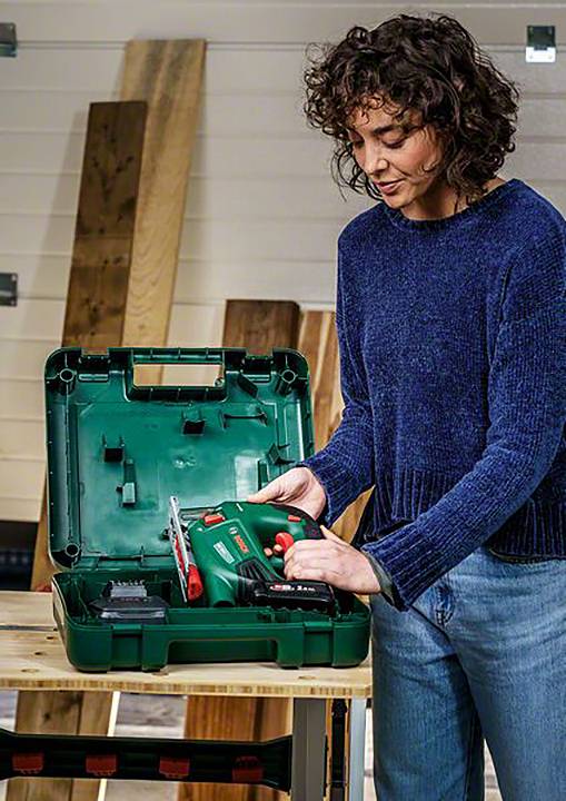 Osoba u puloveru i trapericama stoji kraj stola i drži zelenu bušilicu u otvorenom kovčegu za alat, dok se drvene daske nalaze u pozadini.
