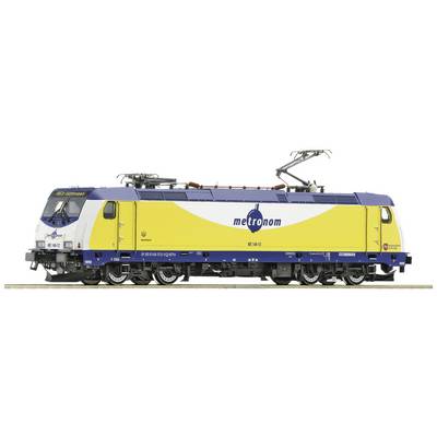 Žuto-plava električna lokomotiva Metronoma s logotipom tvrtke i nadzemnim vodovima. Stoji na dionici željezničke pruge.
