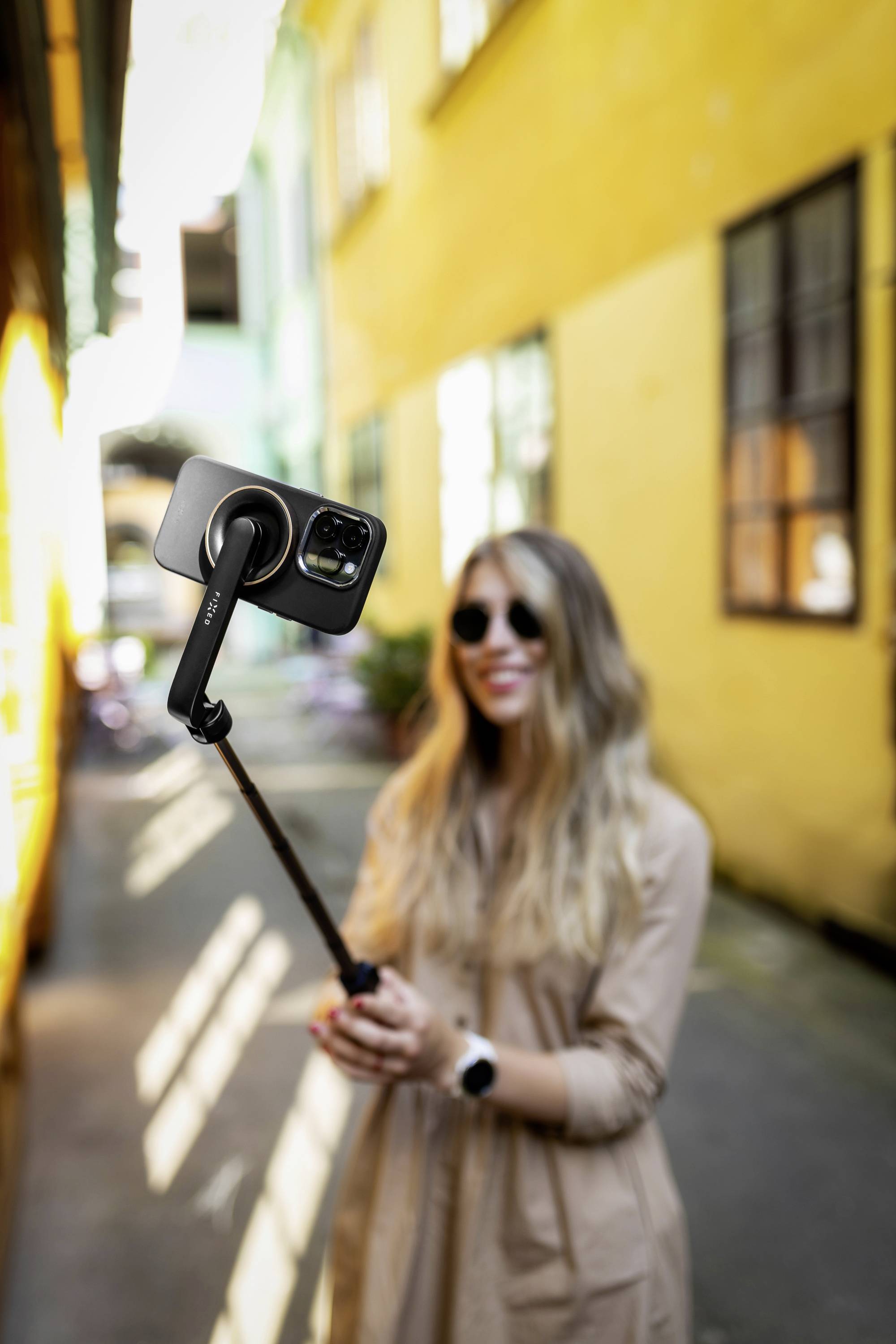 Žena dugih vlasišta drži pametni telefon na selfie štapu. Stoji u sunčanoj uličici s žutim zidovima.
