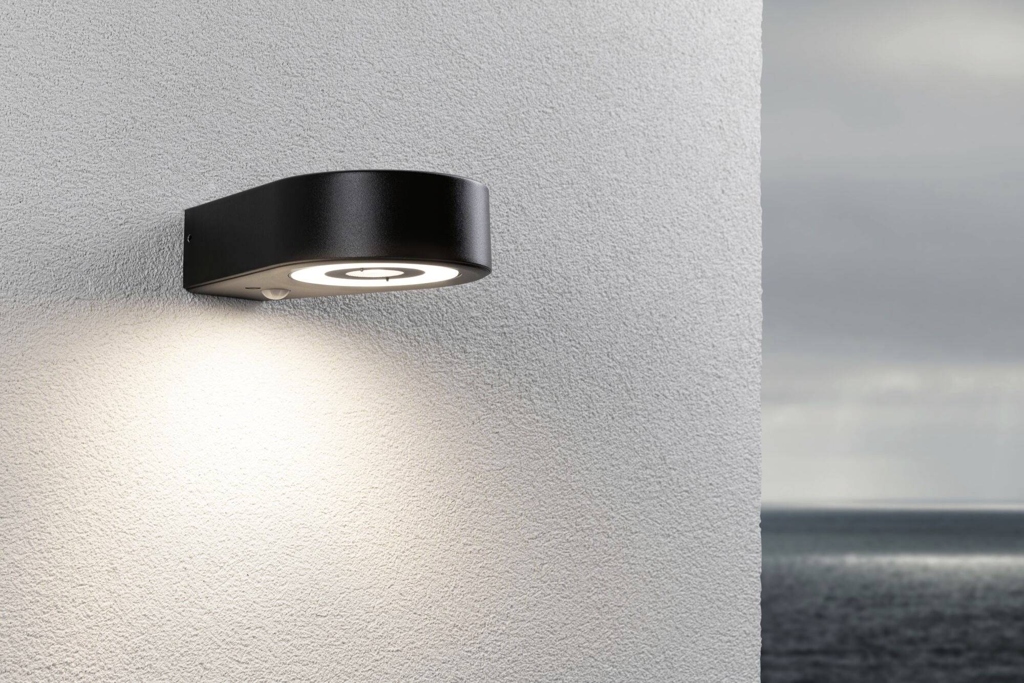 Moderna vanjska zidna lampa osvjetljava bijeli zid; u pozadini se vidi more pod oblačnim nebom.