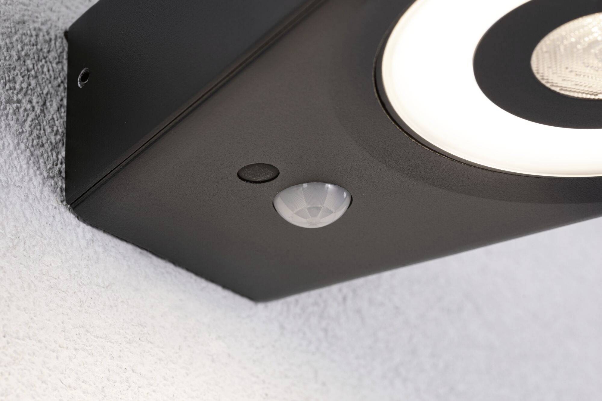 Crna vanjska lampa s senzorom pokreta pričvršćena je na bijelom zidu. Ima okruglo svjetlo na donjoj strani.