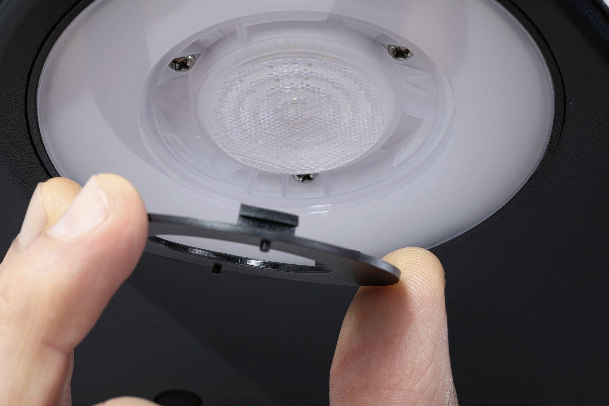 Jedna ruka drži okrugli plastični dio blizu montirane stropne lampe, kao da ga želi postaviti ili ukloniti.