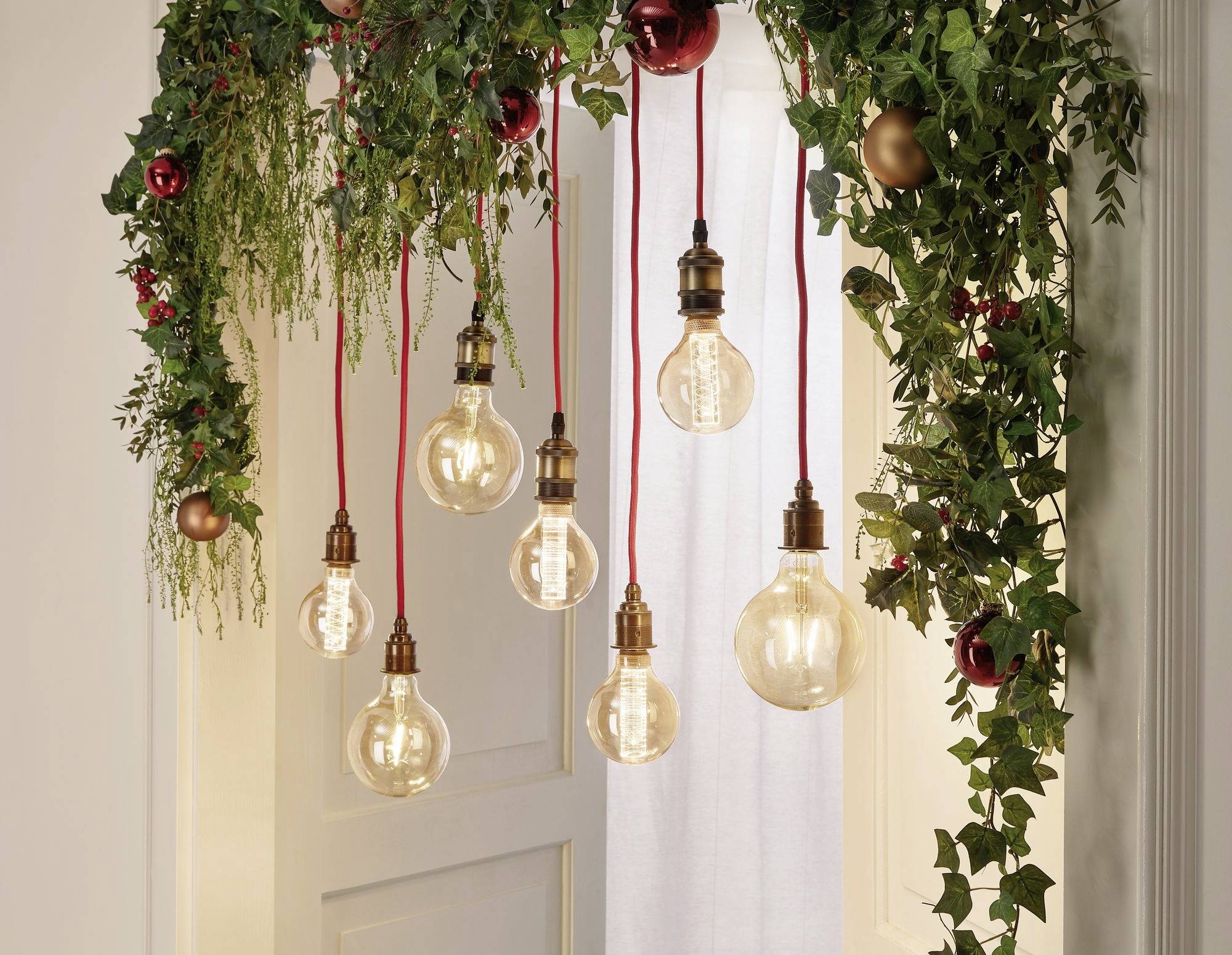 Svečana dekoracija vrata s visećim žaruljama i zelenilom, ukrašena crvenim i zlatnim kuglicama.