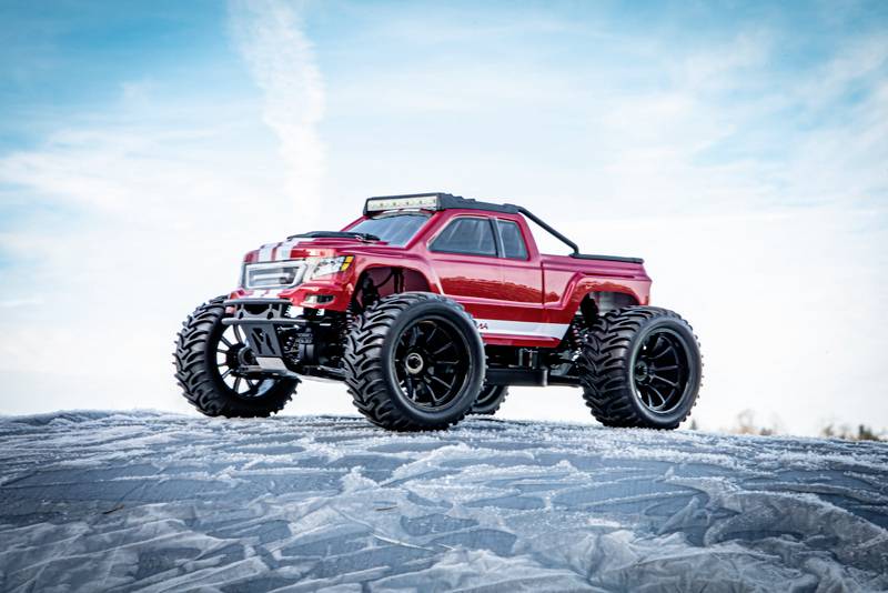 Crveni daljinski upravljani automobil s velikim off-road gumama stoji na snijegom pokrivenom tlu ispred plavog neba.