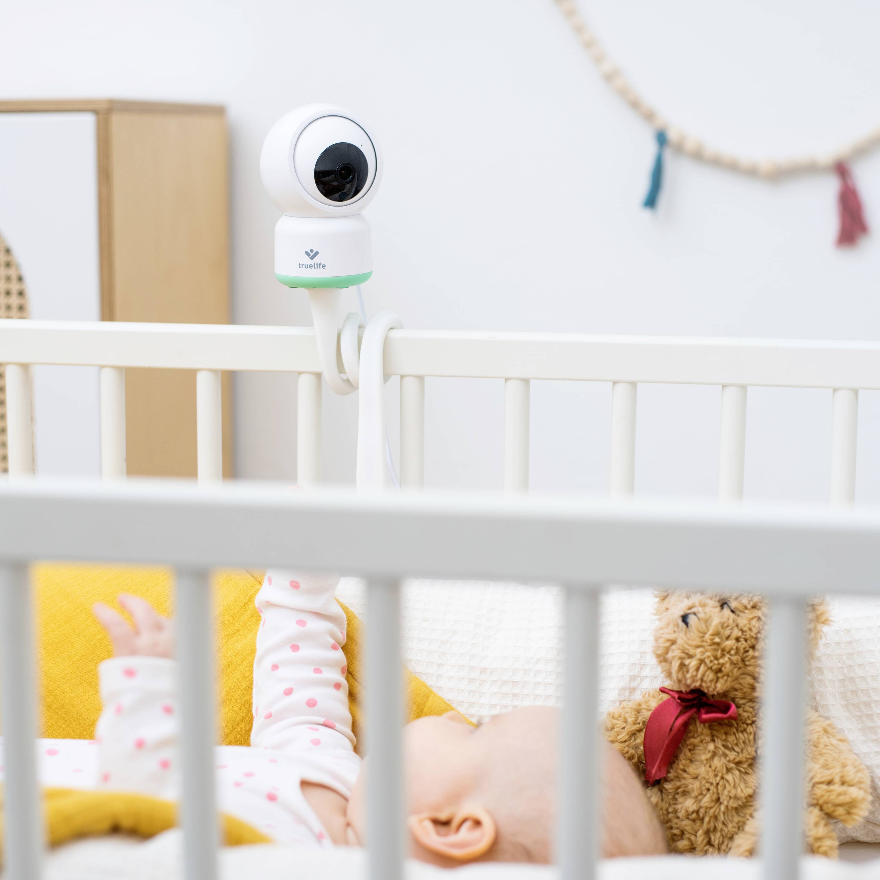 Beba spava u dječjem krevetiću, dok je kamera pričvršćena uz krevet. Plišani medo leži pored bebe.