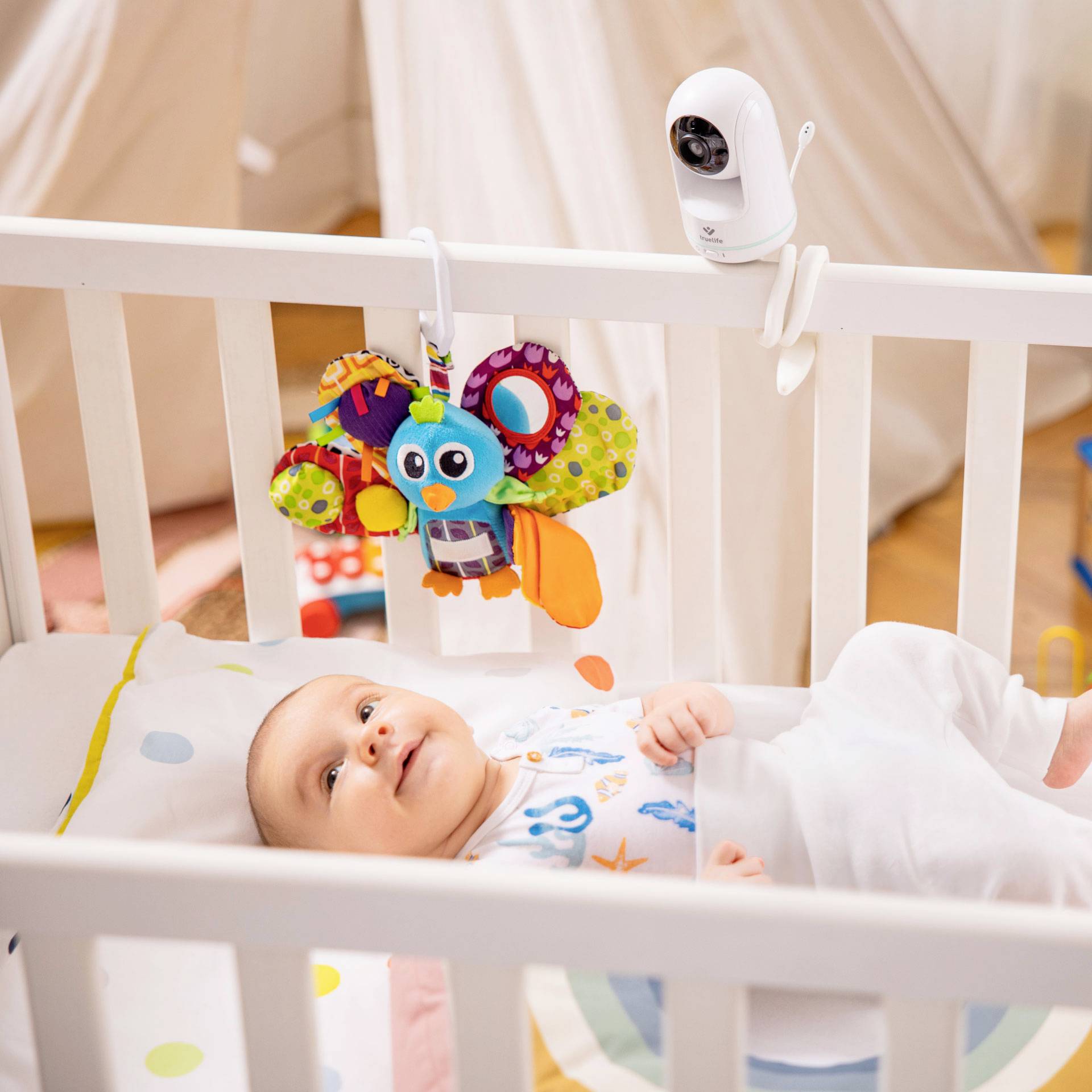 Slika prikazuje malo dijete koje leži u bijelom krevetiću s rešetkama i doseže prema šarenom tekstilnom igračkom. Sigurnosna kamera je postavljena gore na rubu krevetića.