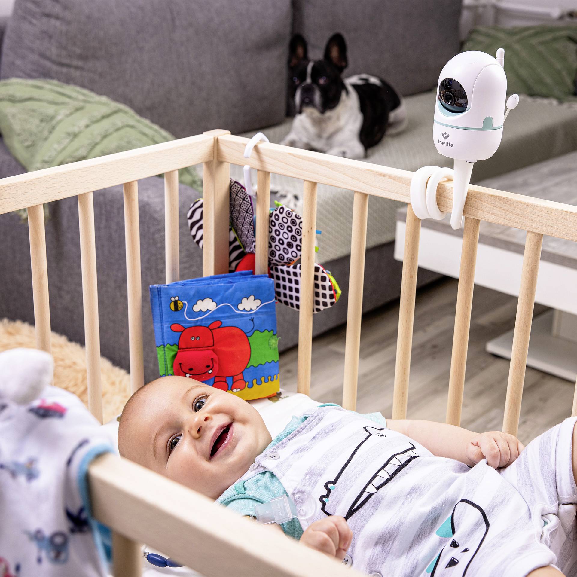 Beba veselo leži u krevetiću za djecu okružena igračkama. Kamera je pričvršćena za rub kreveta. Mali pas sjedi u pozadini.