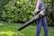 Osoba puše lišće u vrtu pomoću leaf blowera. Pozadina prikazuje zelene biljke. Široka travnata površina u prednjem planu.