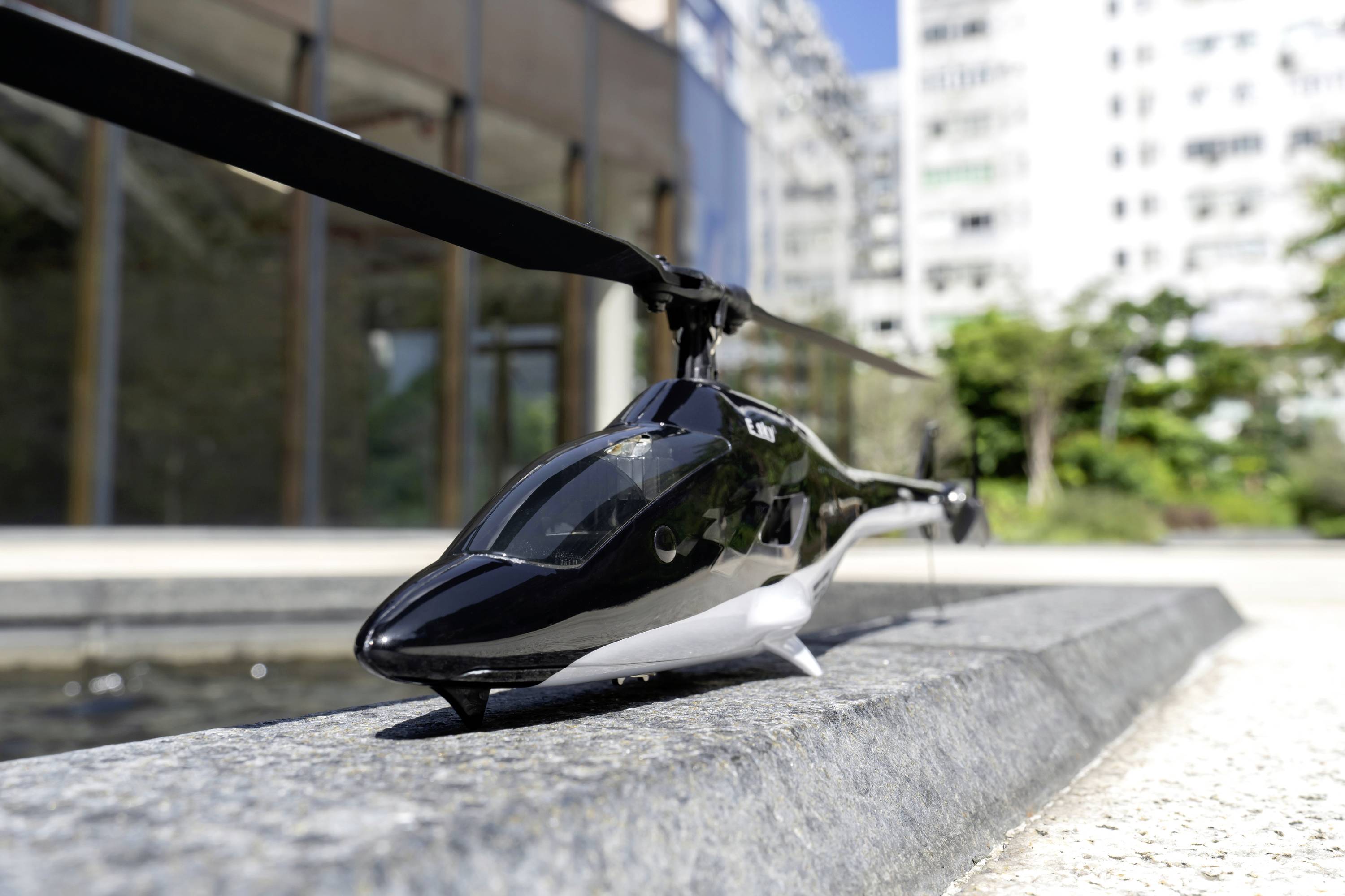 Modelarski helikopter stoji na rubu pločnika, okružen modernim zgradama i zelenim površinama.
