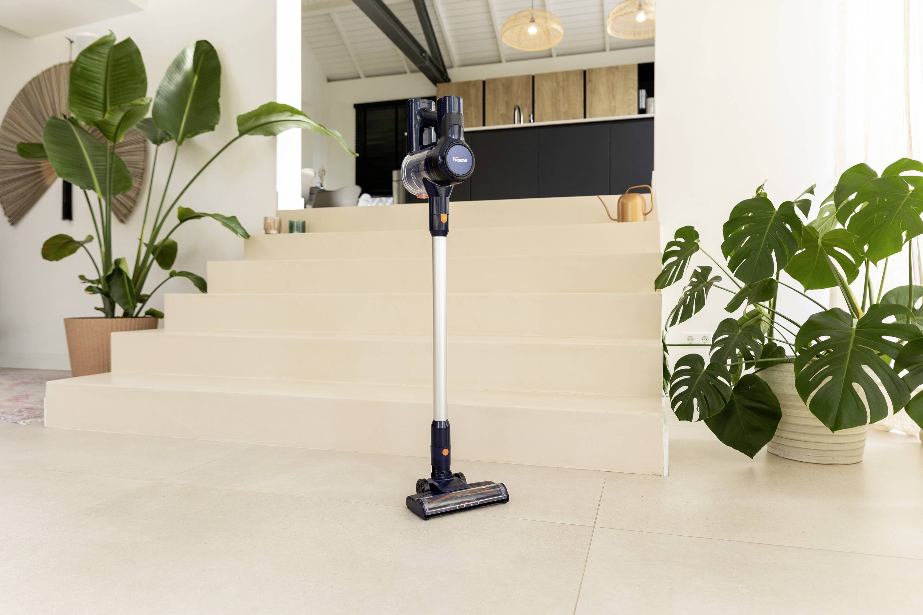 Bežični usisavač stoji na svijetloj podlozi ispred stubišta u moderno uređenoj prostoriji s velikim sobnim biljkama.