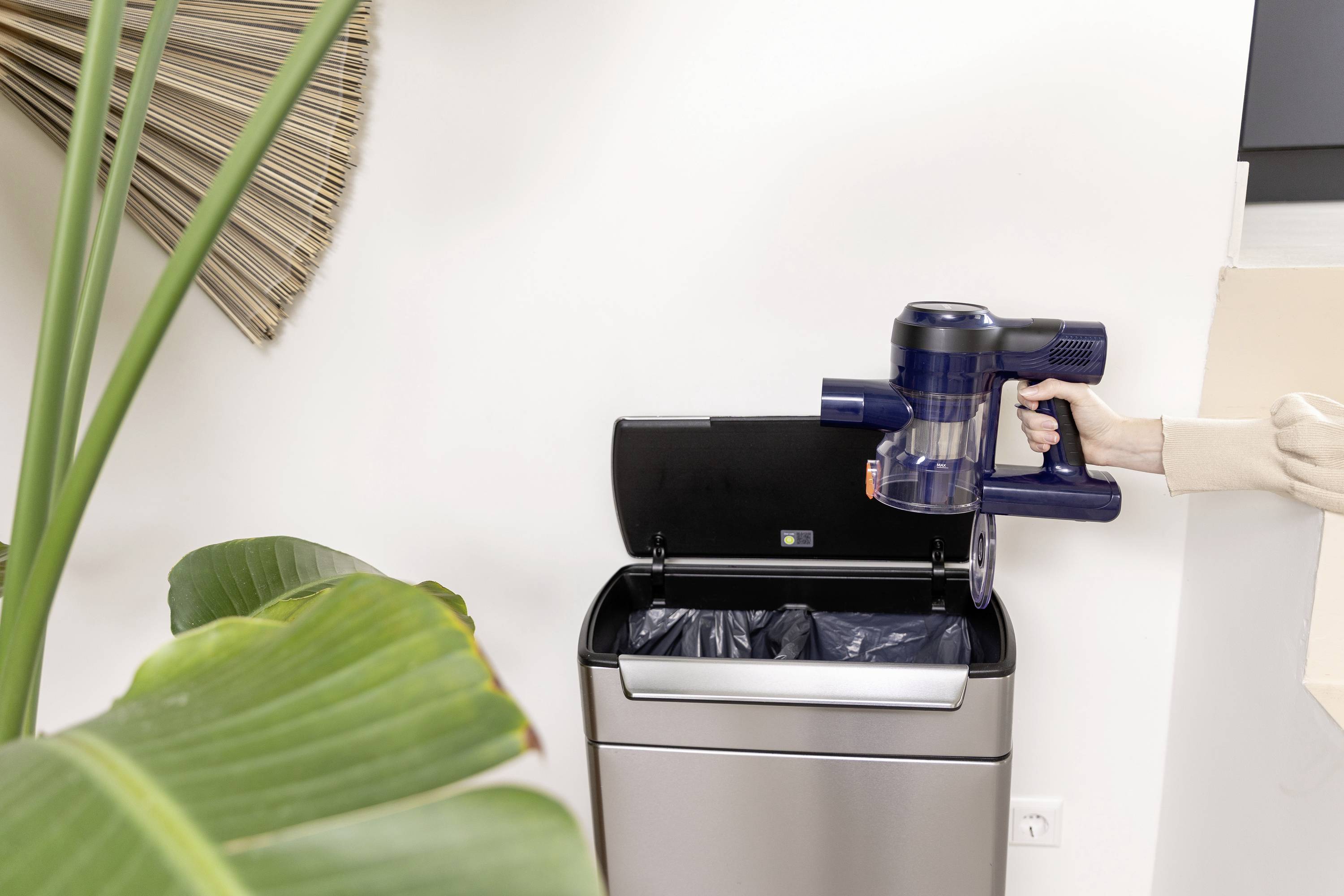 Osoba prazni spremnik bežičnog usisavača u košu za smeće. U pozadini sobna biljka i moderna unutrašnjost.