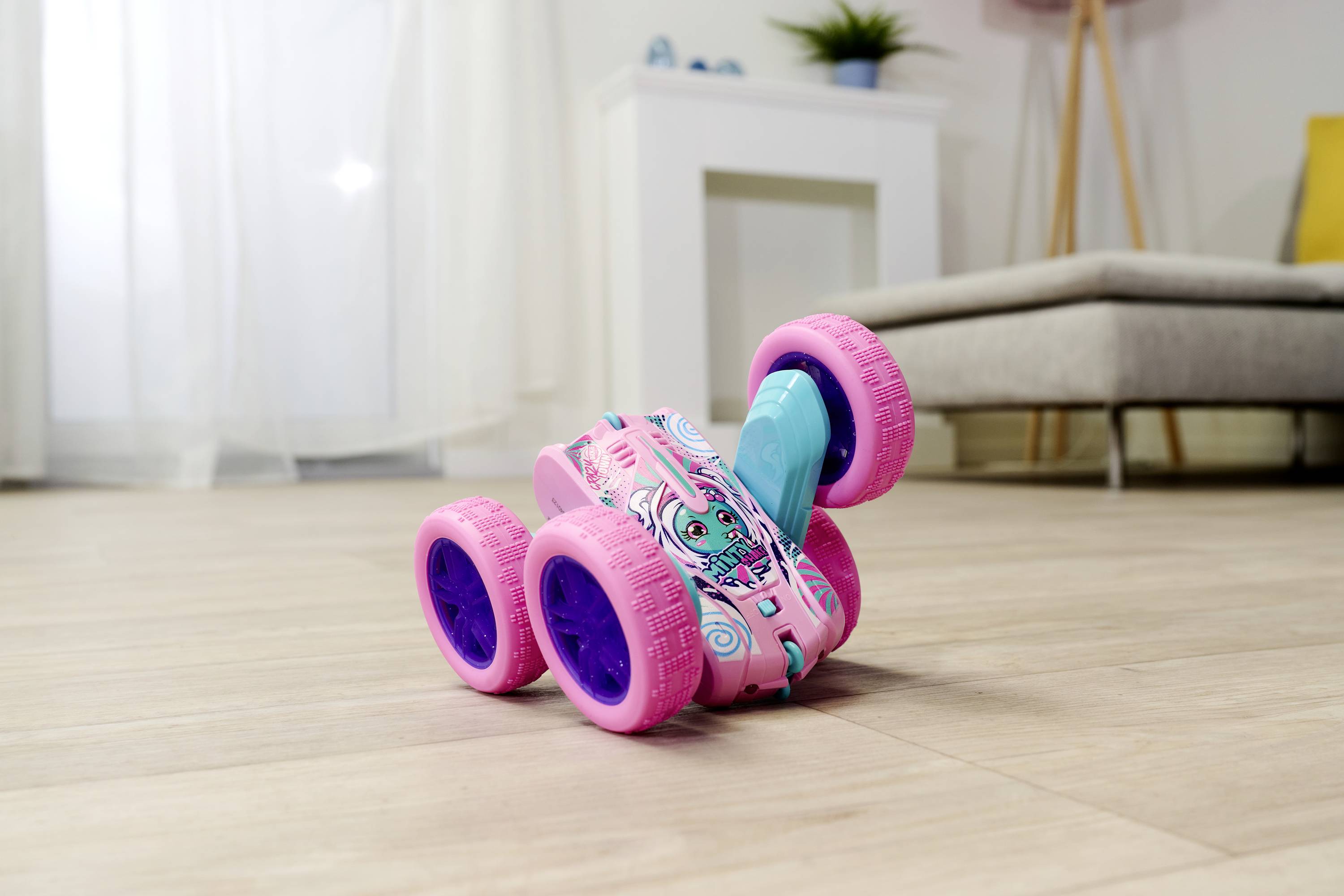 Ružičasto-plavi igračka automobil s četiri velika kotača stoji na drvenom podu u dnevnoj sobi, u pozadini je sofa i biljke.